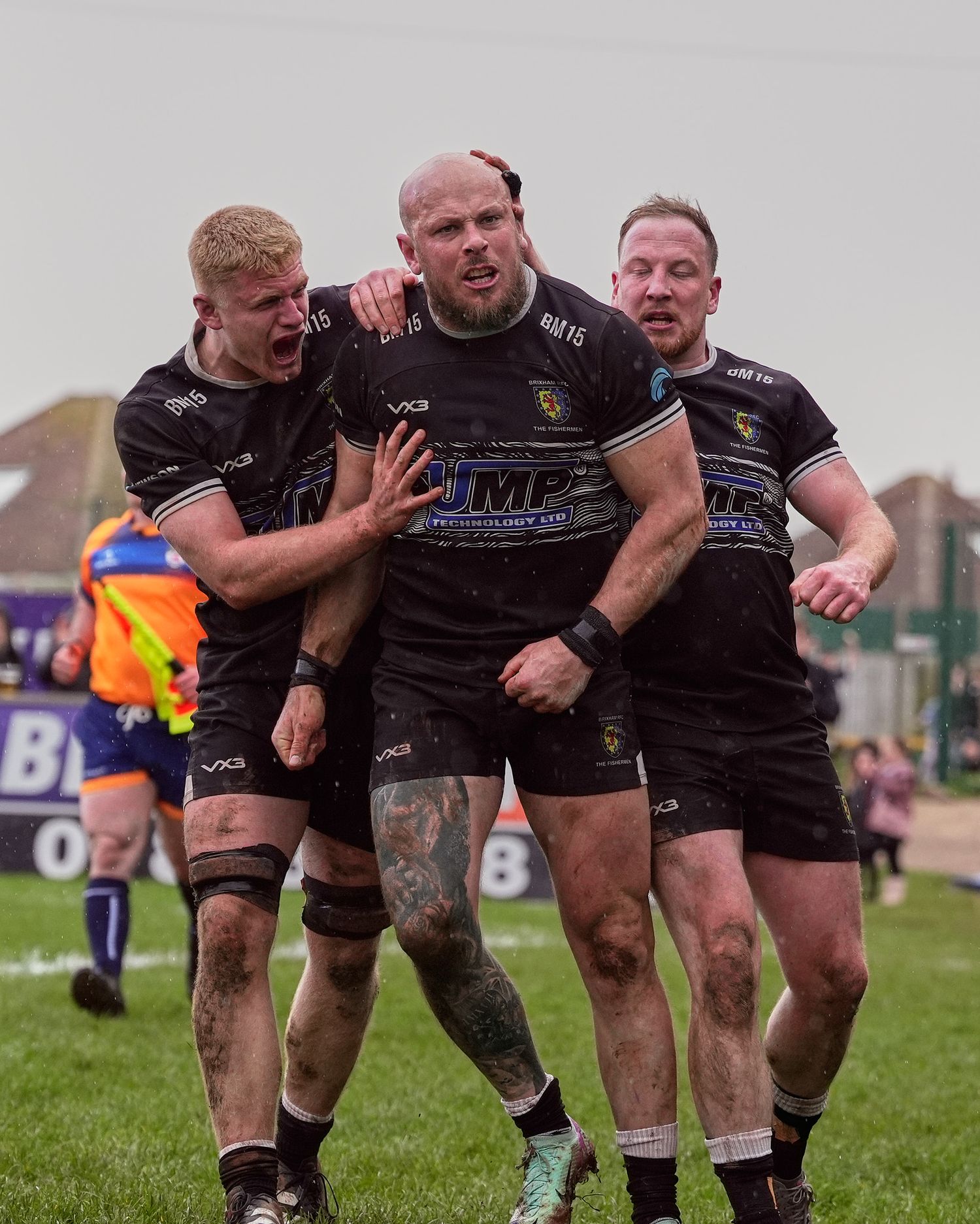Three Brixham RFC rugby players in black kit celebrate together on a muddy field during a match.