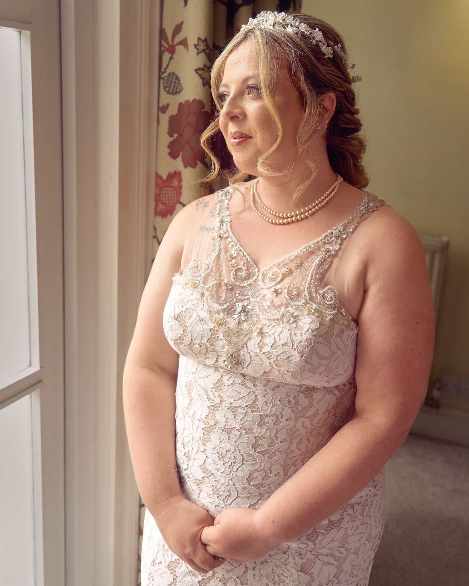 Bride in a lace wedding dress standing beside a window for a quiet bridal portrait.