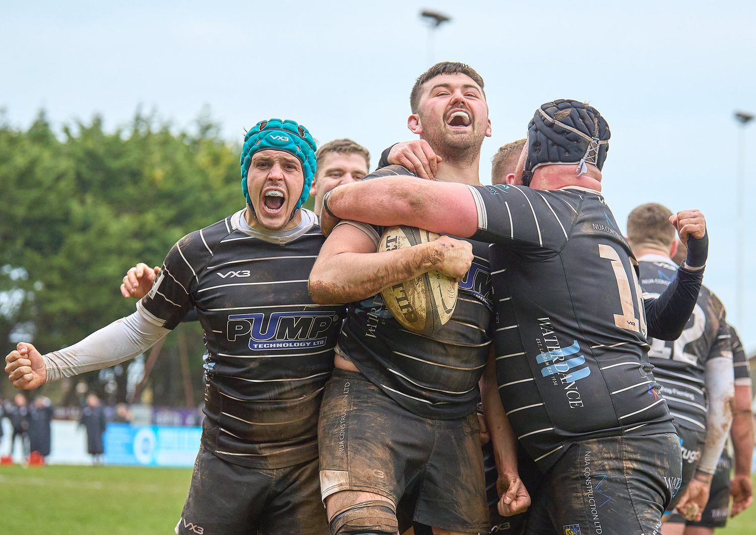 Brixham RFC players celebrate a try in muddy match, wearing dark jerseys.