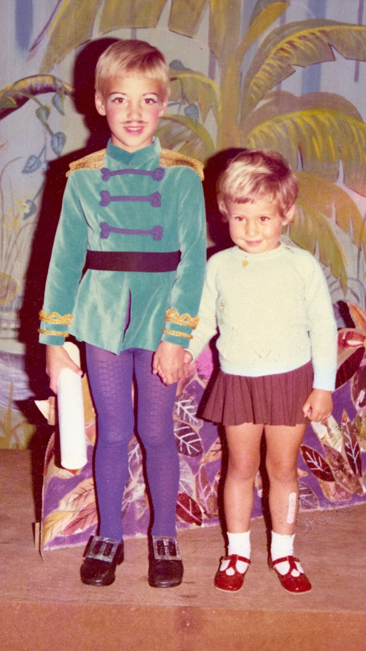 Two children stand together, one wearing 1970s style clothing, the other dressed as the prince from Cinderella against a painted backdrop.