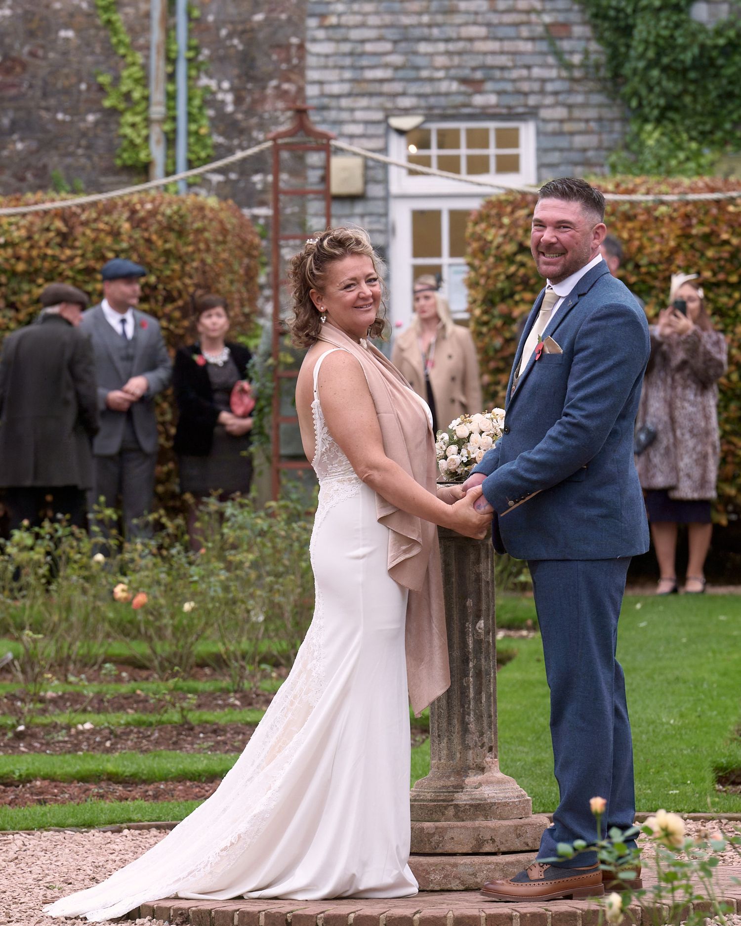 Bride and groom standing together outdoors with wedding guests behind them.
