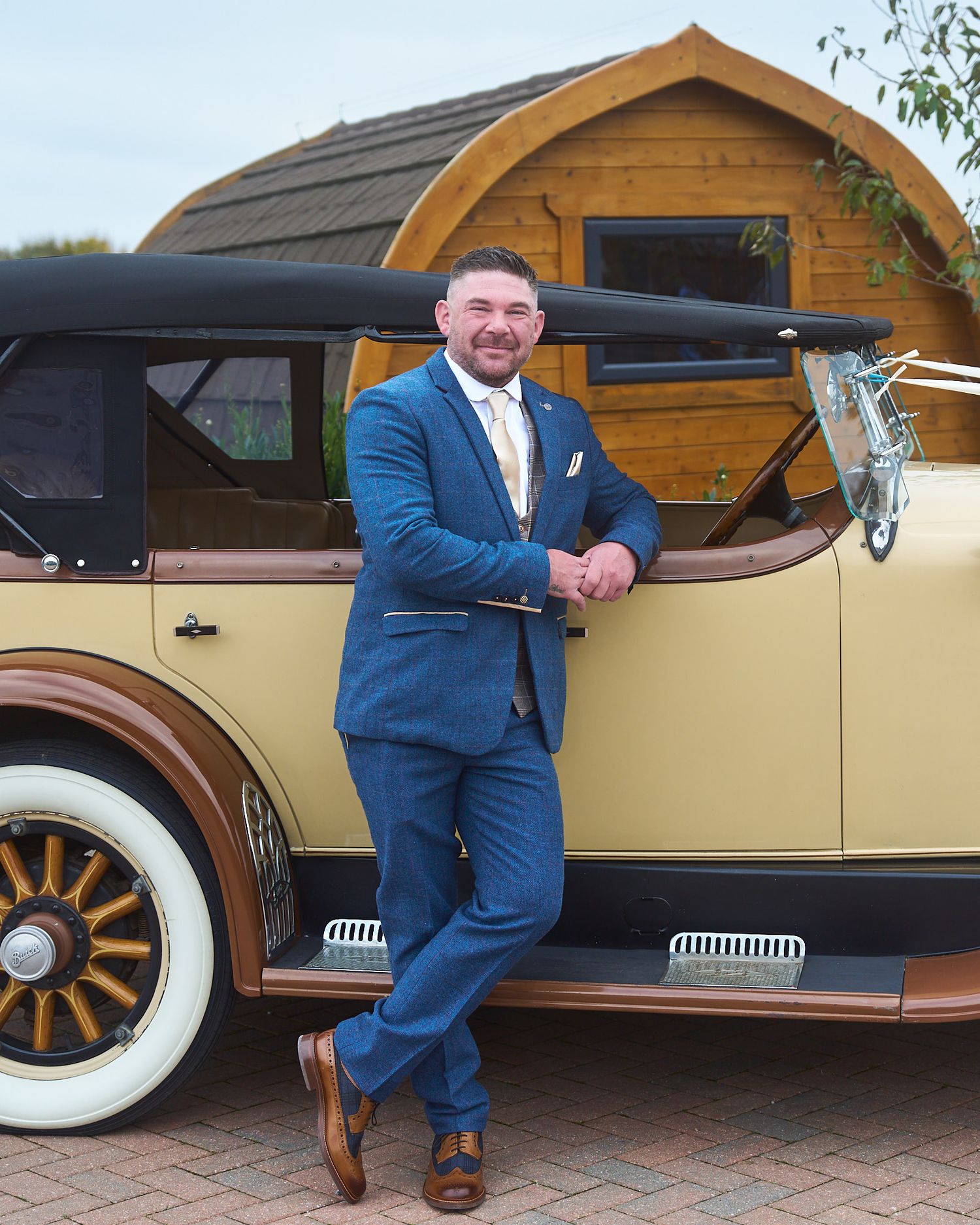 Groom in a blue suit posing beside a vintage wedding car before the ceremony.