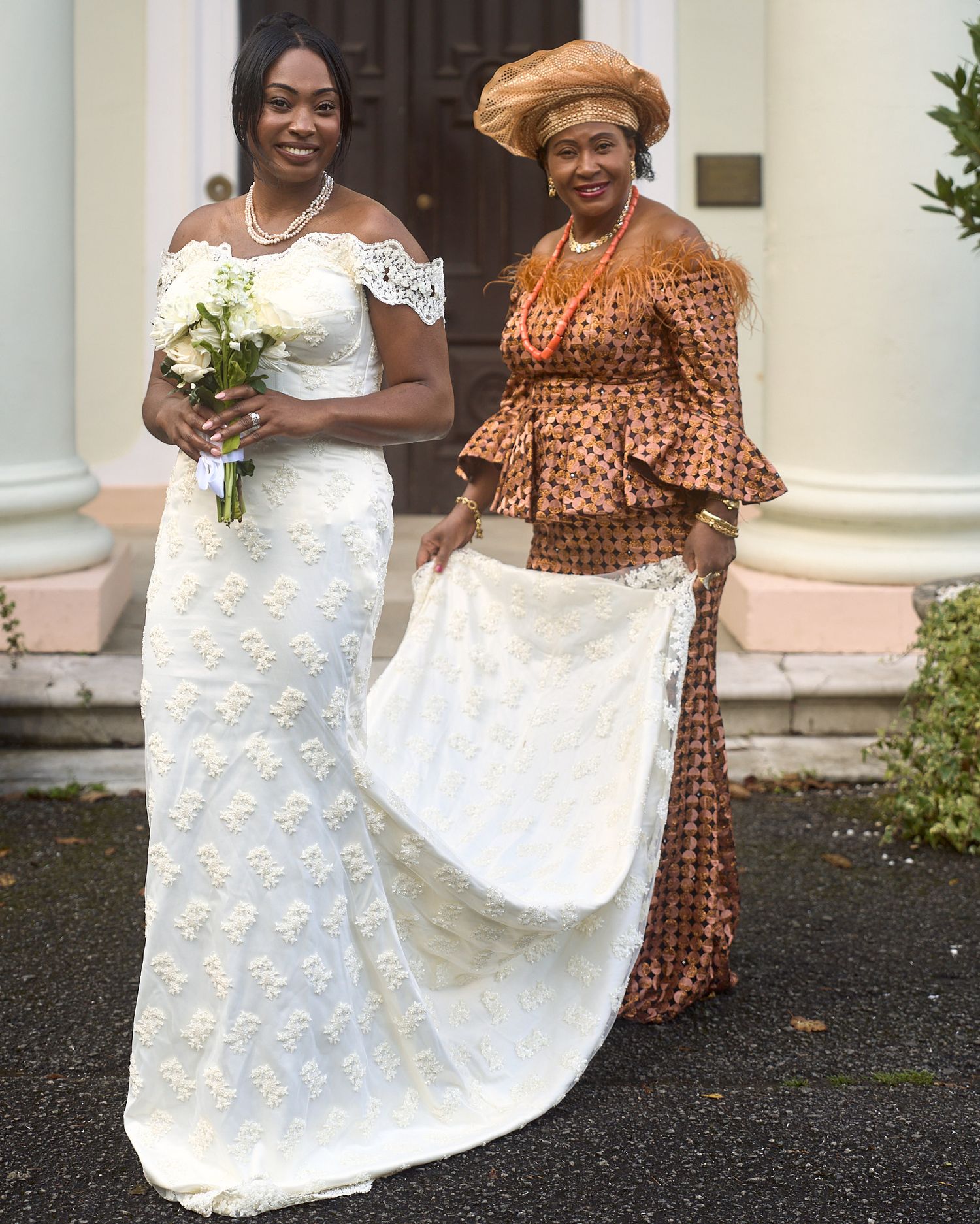 Bride in a lace gown with a woman in traditional attire helping hold her wedding train.