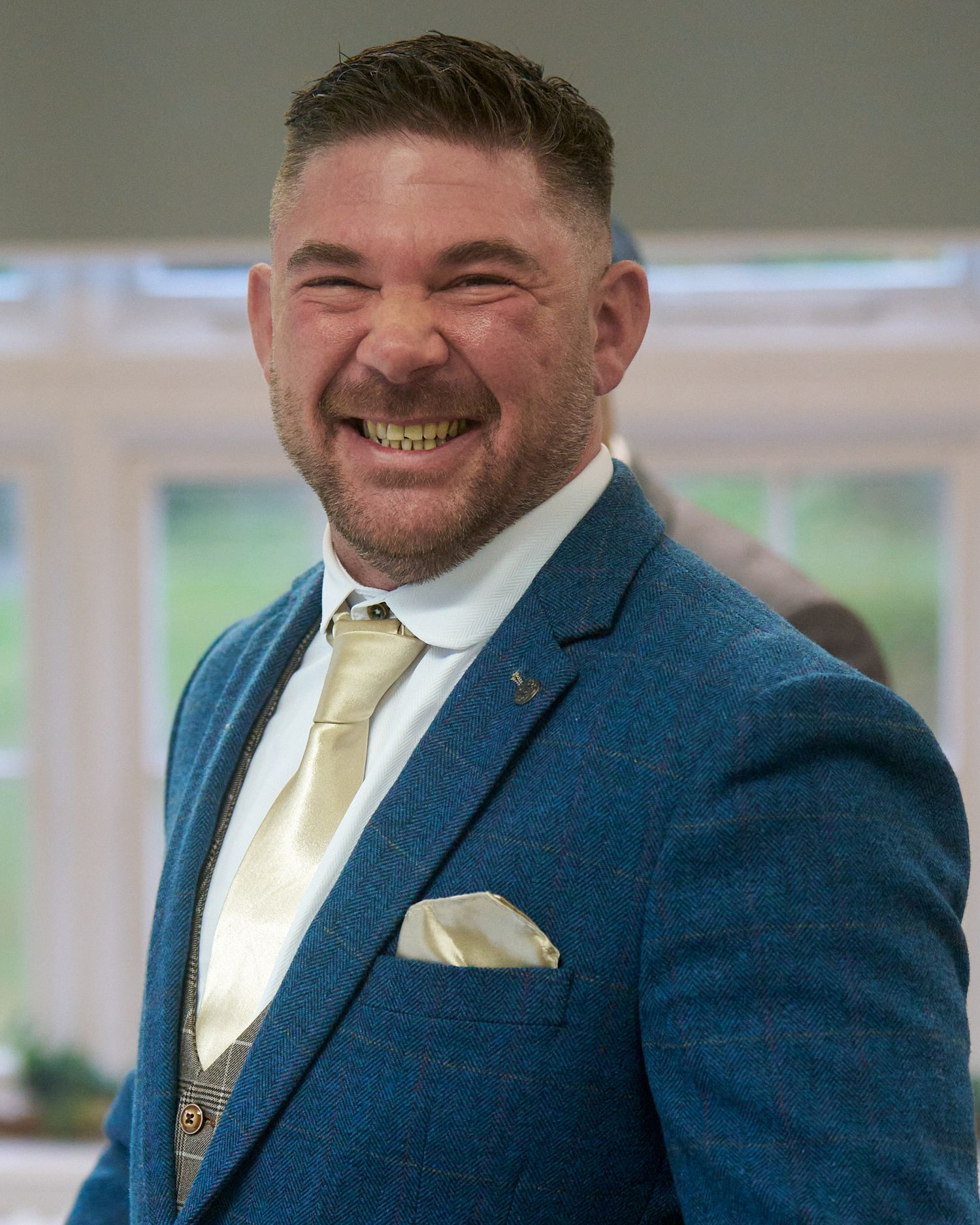 Smiling groom in a blue suit and cream tie during indoor wedding preparations.