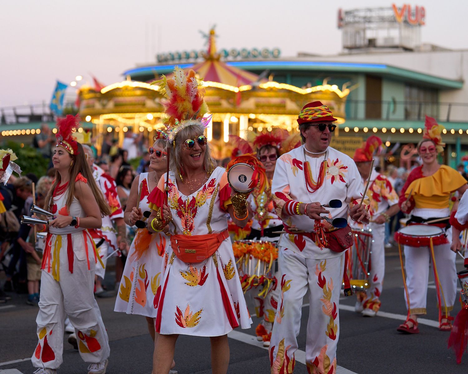 Colorful carnival parade performers in white and red costumes marching past a glowing carousel at dusk.