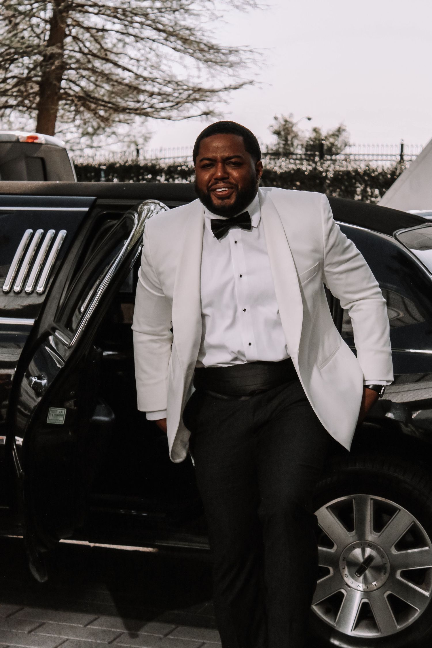Wedding party members in formal white tuxedo jackets pose beside black limousine.