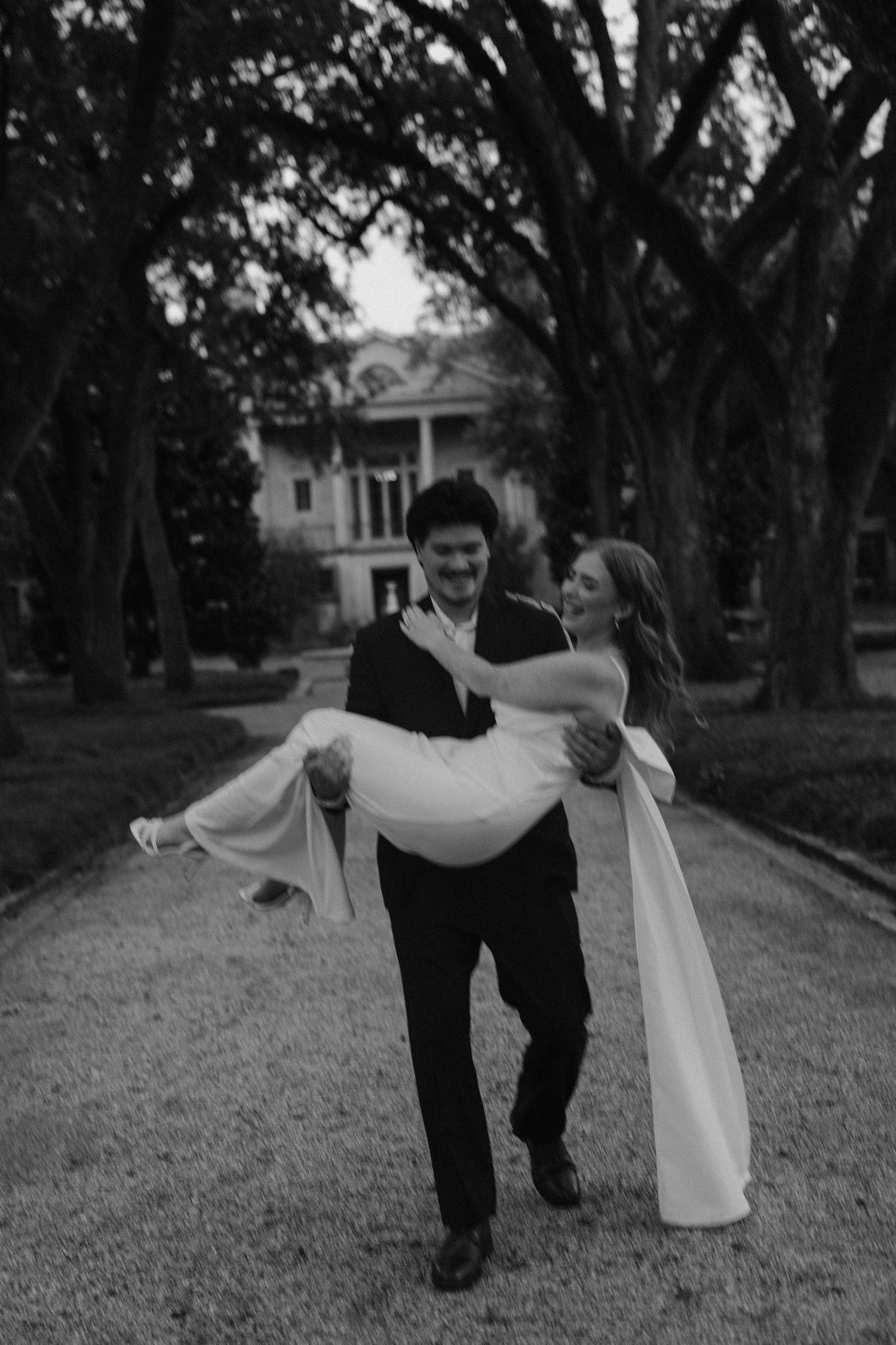 Groom carries bride in white dress along tree-lined path with historic mansion in background, black and white wedding photo.