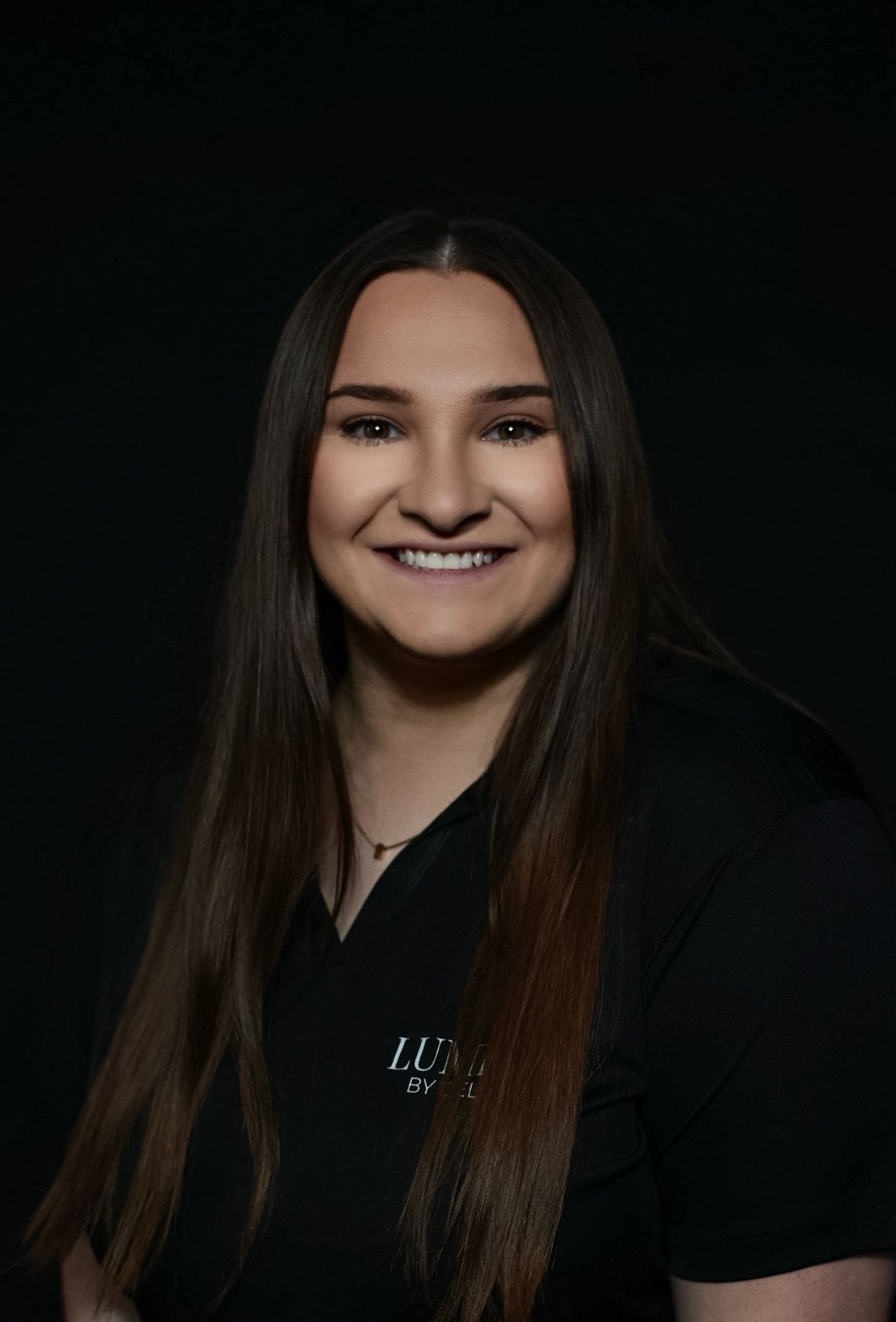 Young woman with long brown hair smiling, wearing a black Lumi branded shirt against a dark background.
