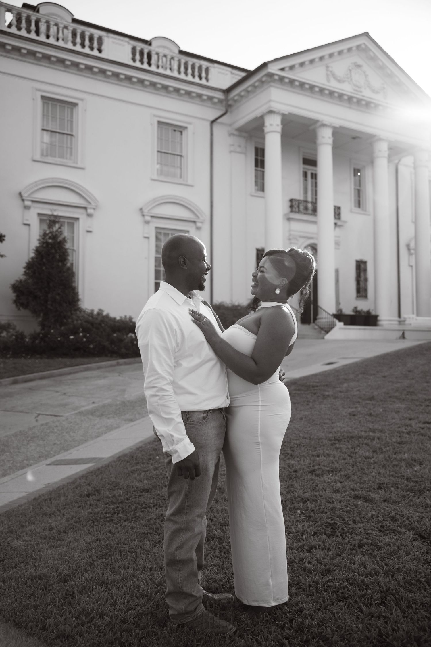 A romantic couple shares an intimate moment on the lawn in front of a grand white columned mansion.