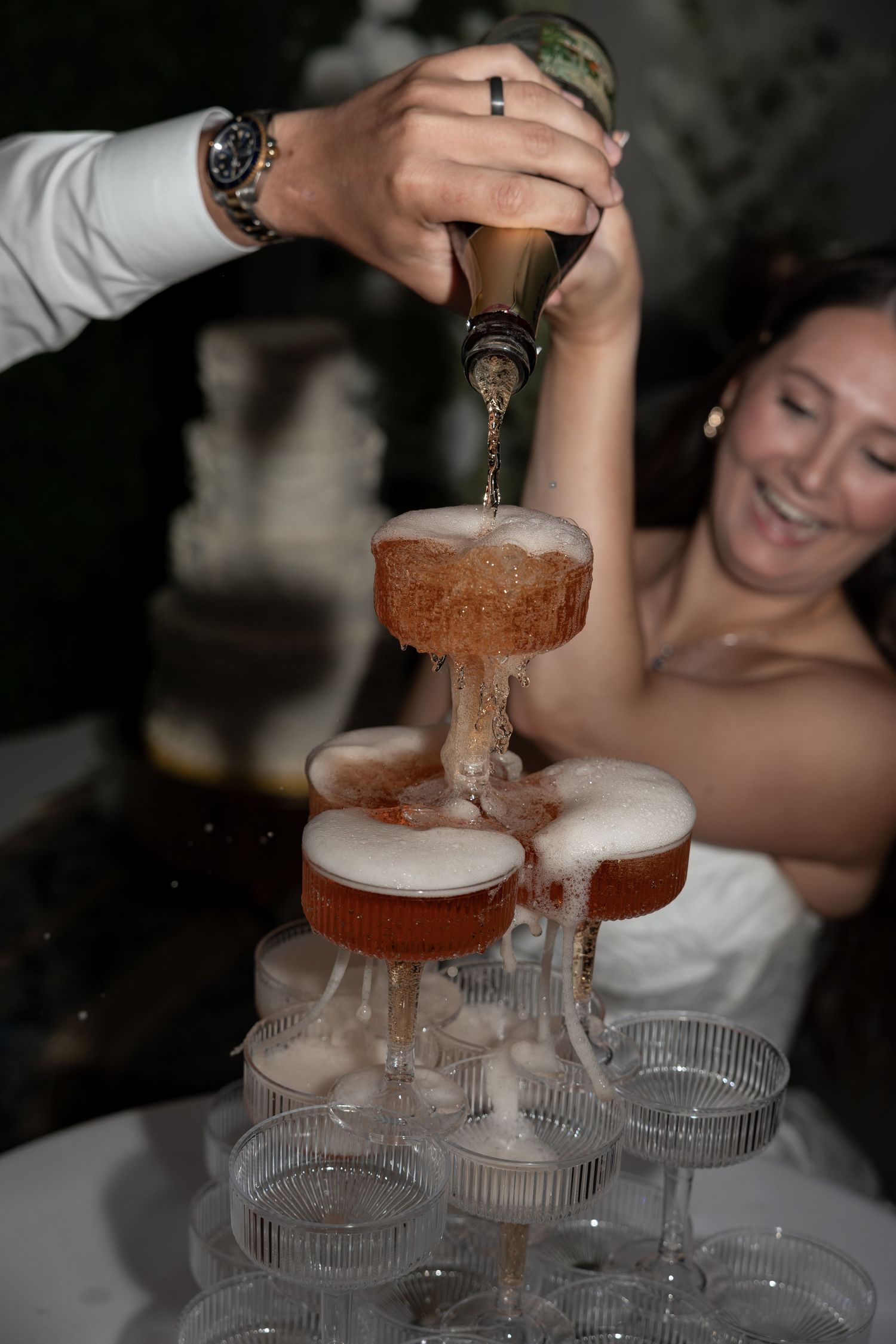 Bride smiles as groom pours champagne over a glass tower at their wedding reception celebration.