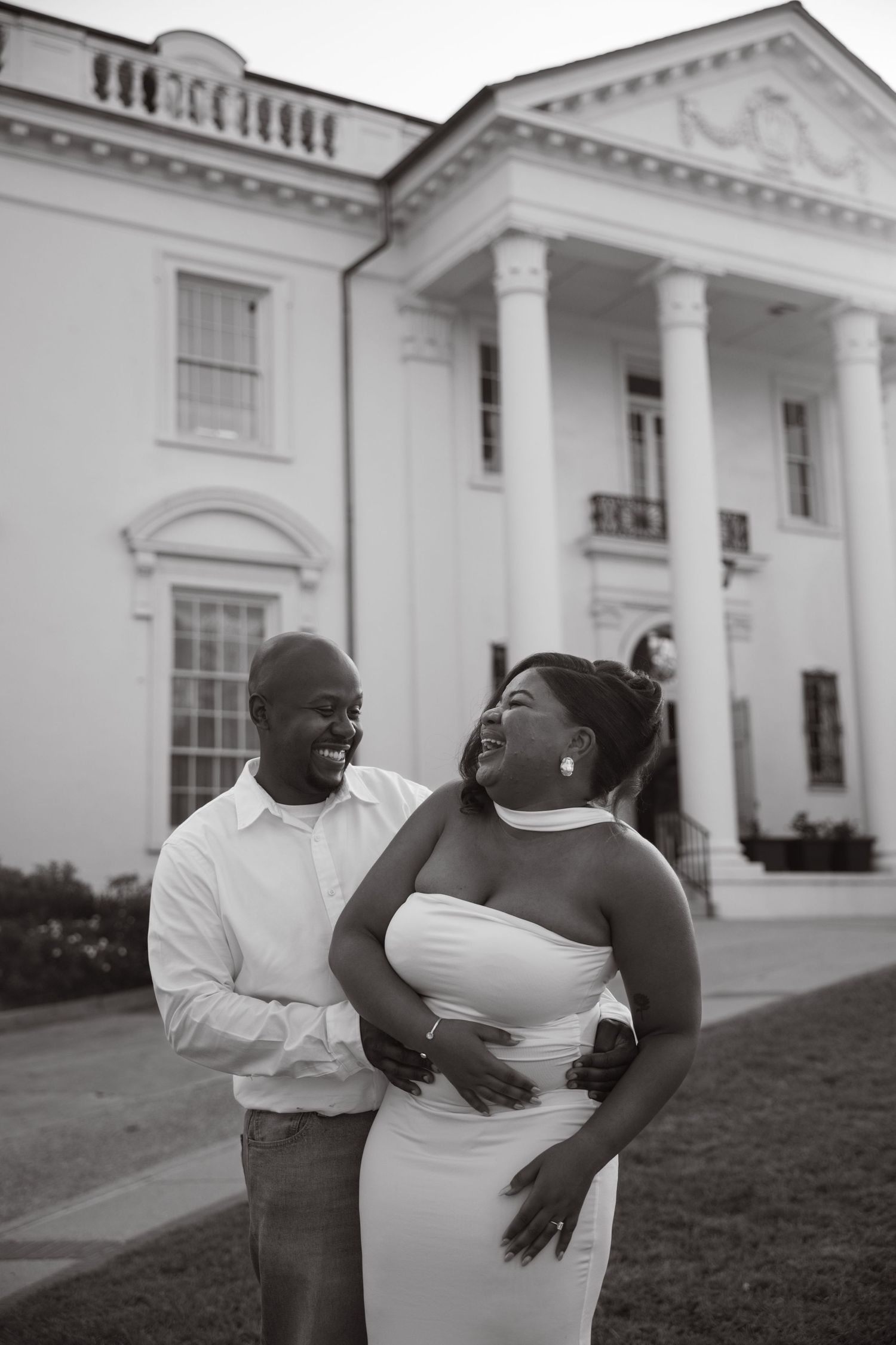 Black and white photo of a joyful couple laughing together in front of a grand white columned mansion.