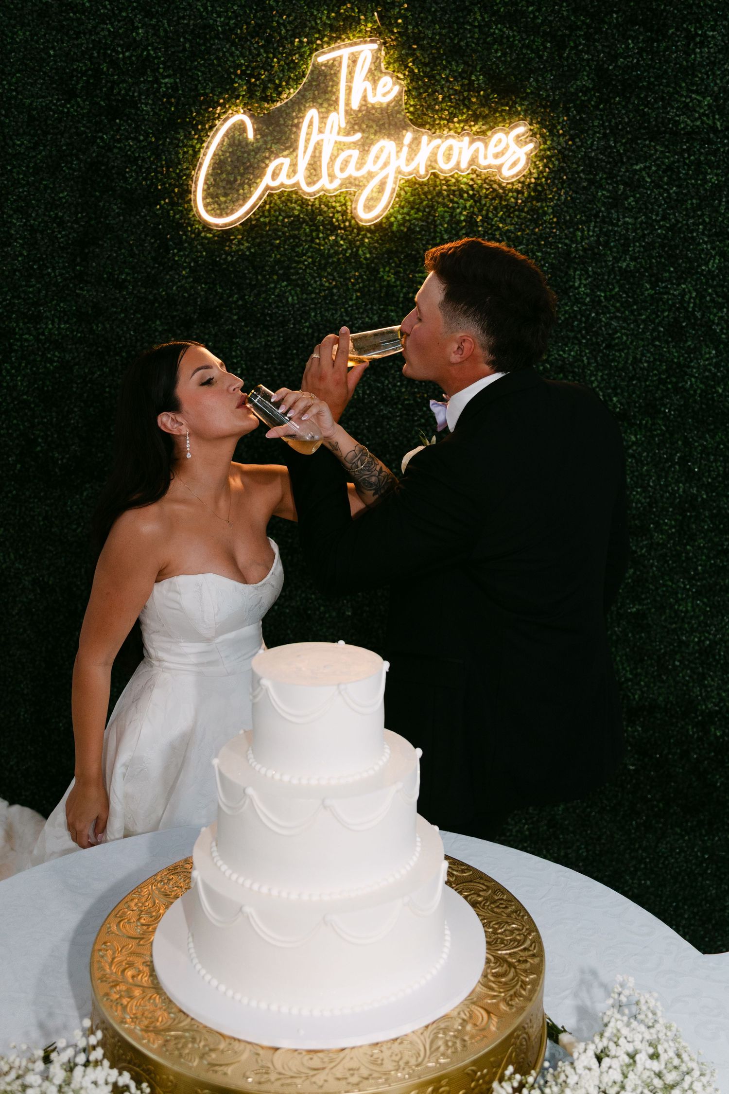 Bride and groom toasting champagne at wedding reception with white tiered cake and neon sign reading The Callagirones.