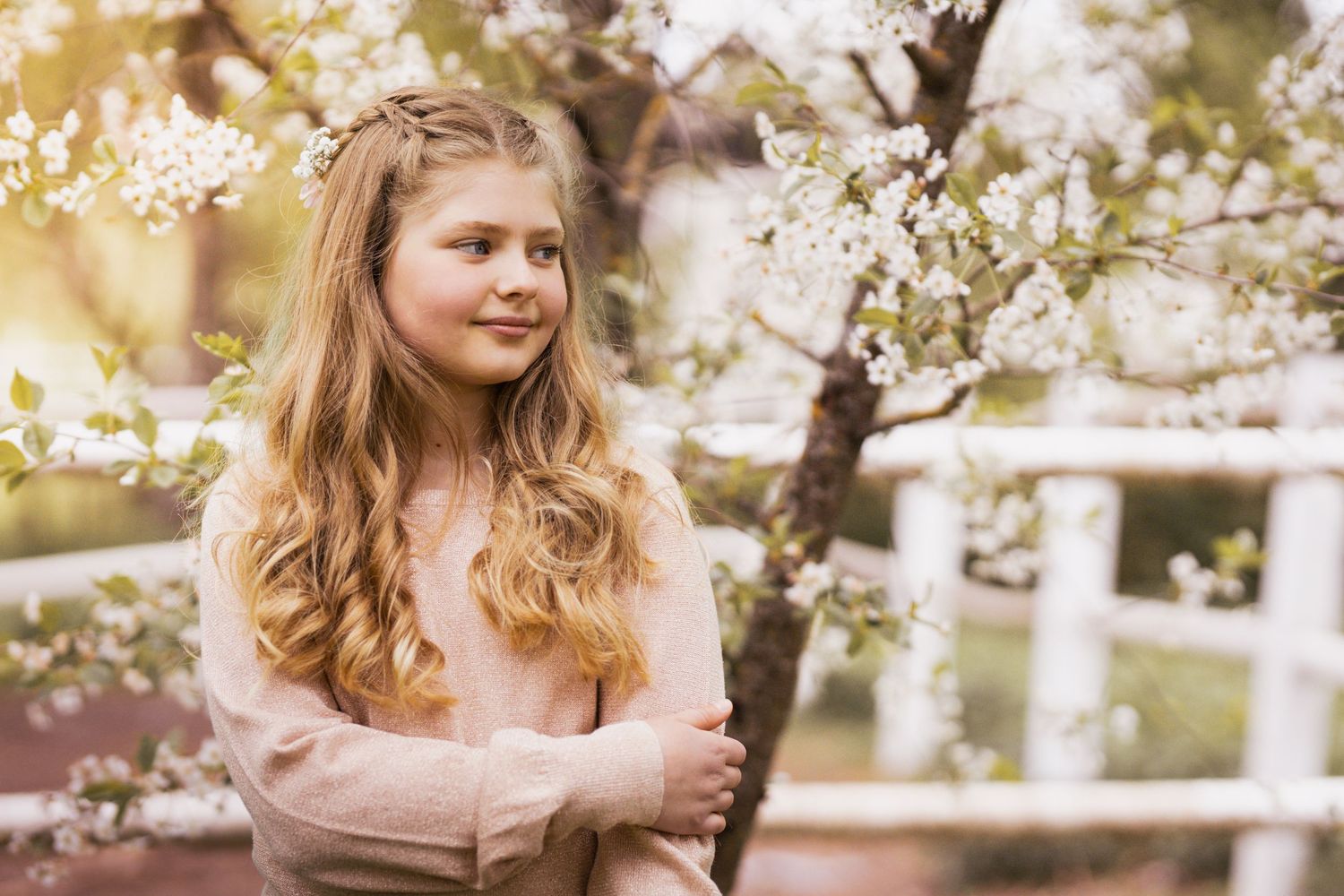 A person with long wavy hair poses near blooming spring cherry blossoms in a soft pink sweater.