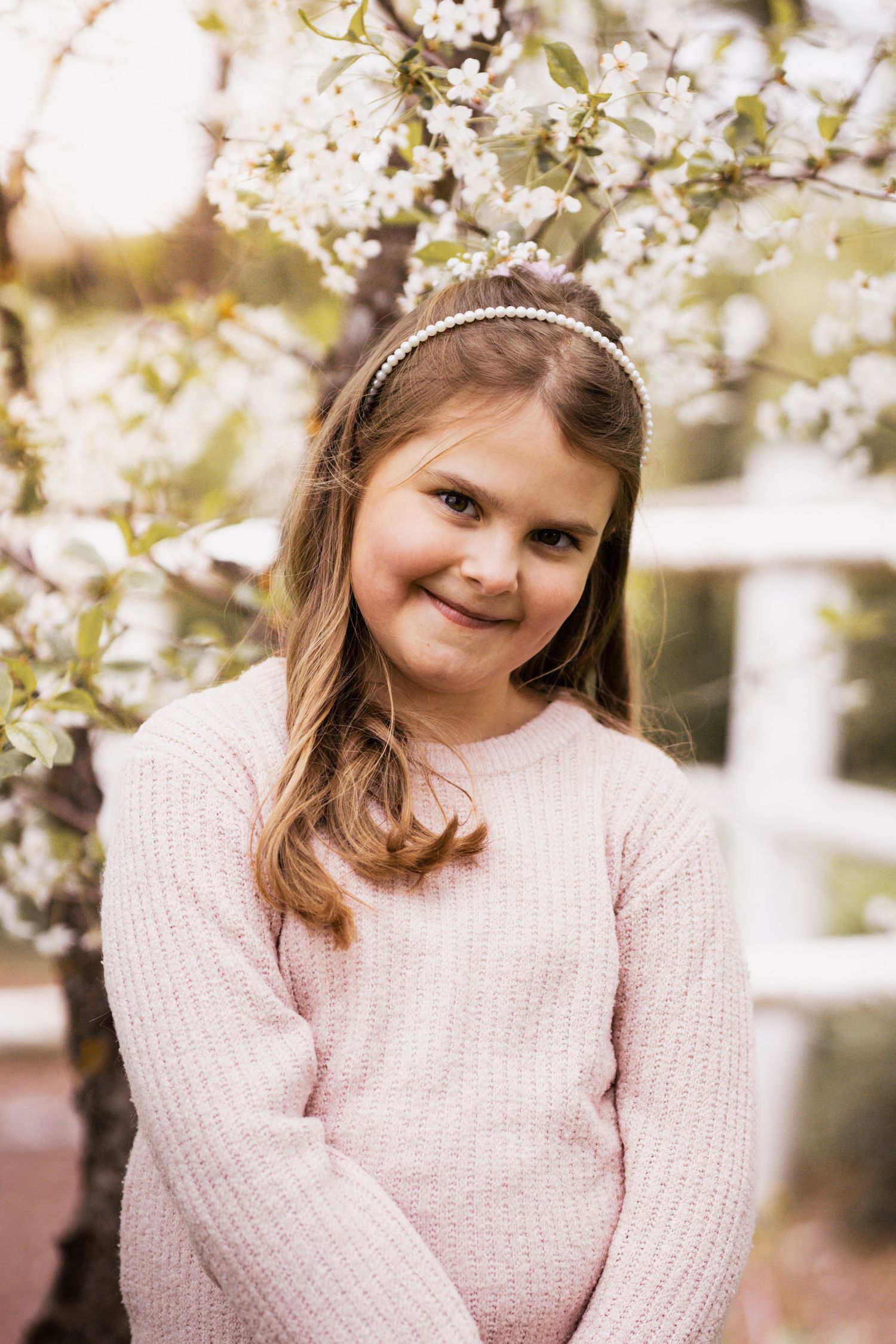 A young girl in a pink sweater smiles warmly while posing near blooming cherry blossoms in springtime.