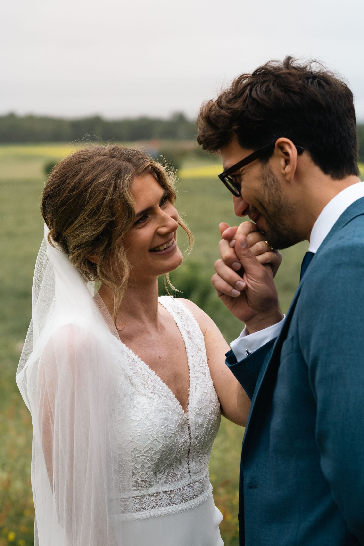 A bride in a lace wedding dress and groom in blue suit share intimate moments in a field.