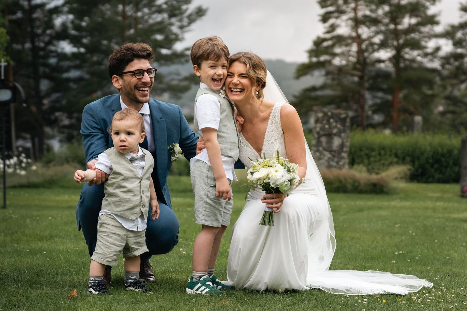 A playful wedding photoshoot in a green outdoor setting with pine trees in the background.