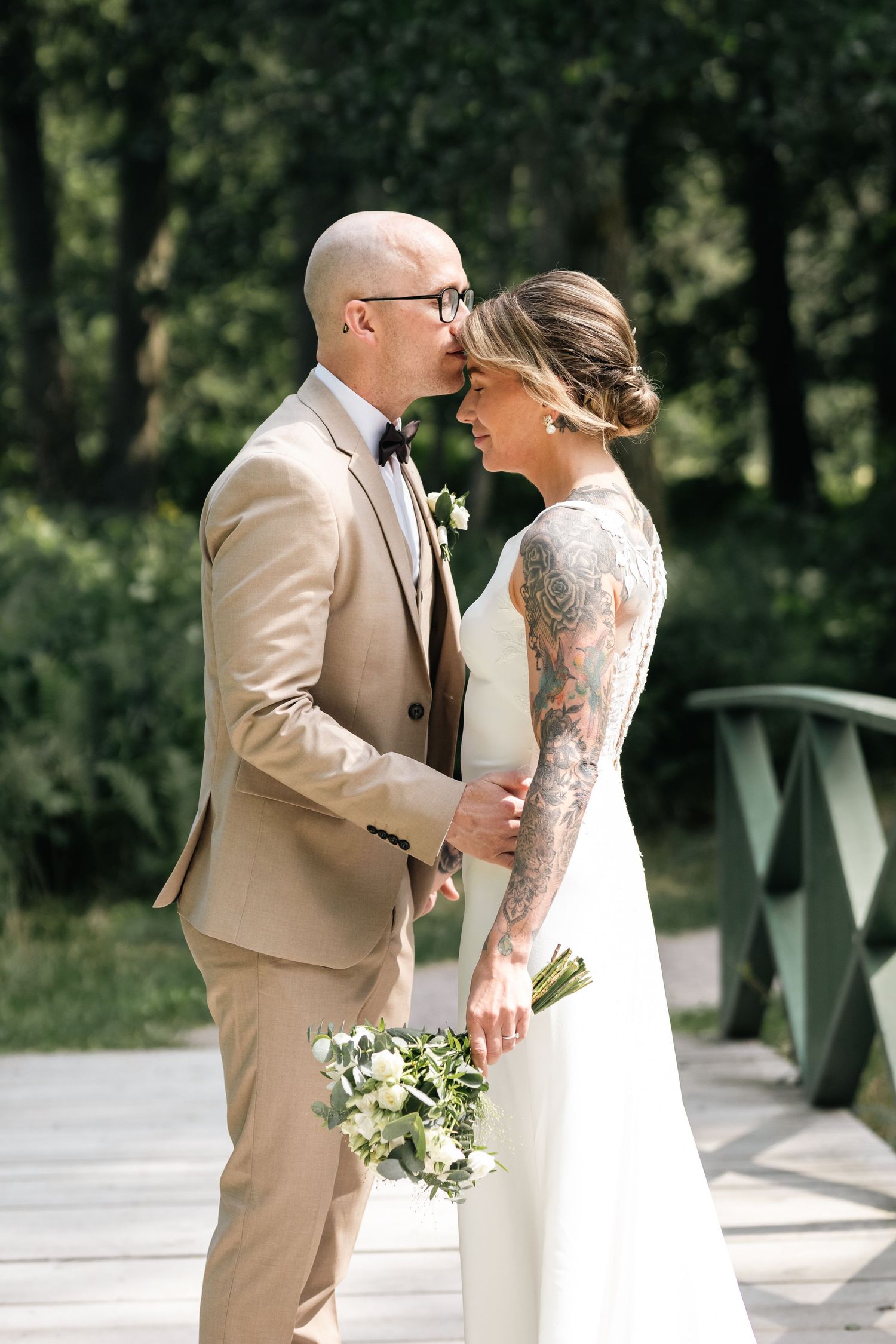 A wedding couple in tan and white attire share an emotional first look moment on a bridge.