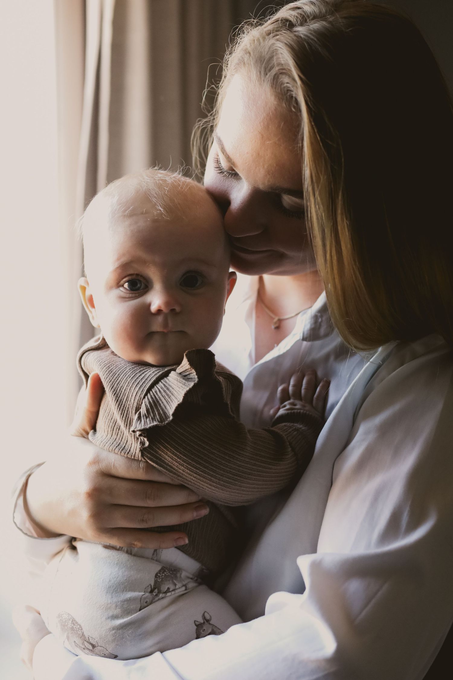 Mother and baby share tender moment in soft window light.