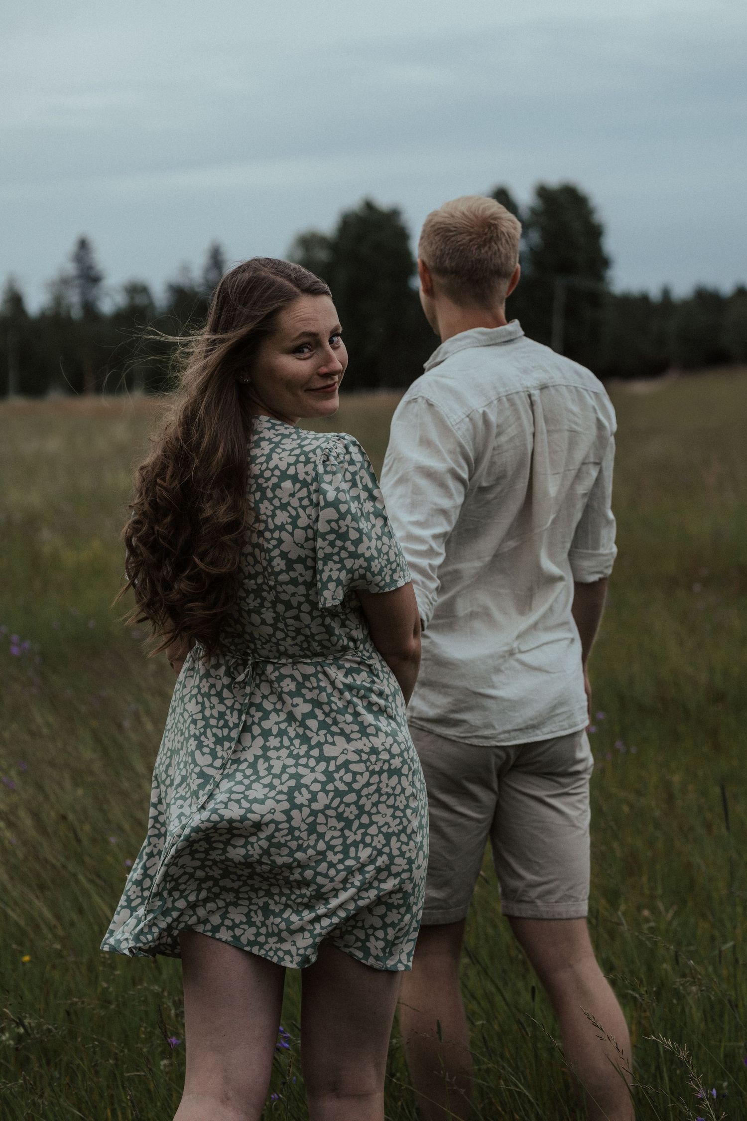 A playful couple running through a grassy field on a cloudy day.