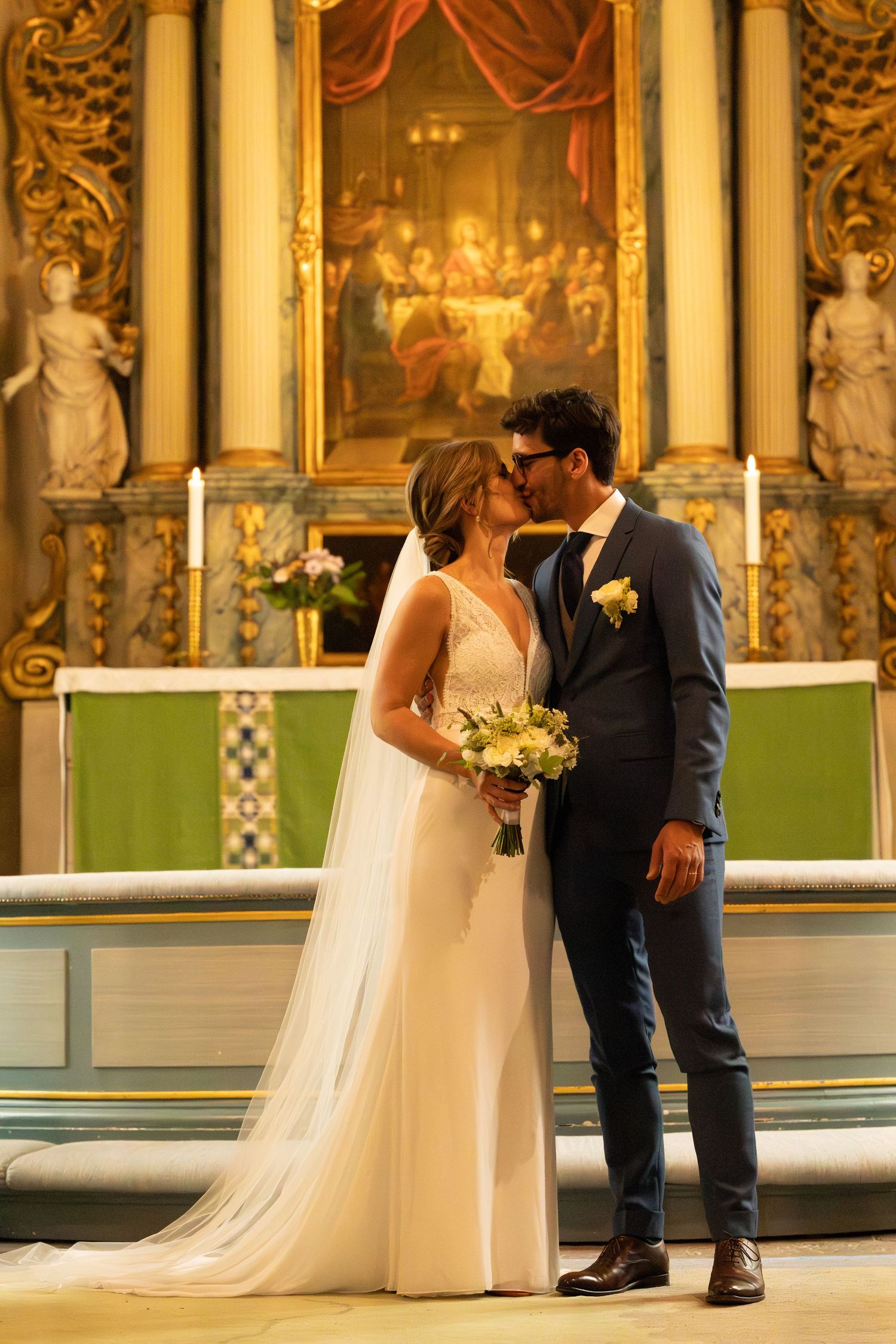 A wedding couple shares an intimate moment in an ornate church interior with golden columns and paintings.