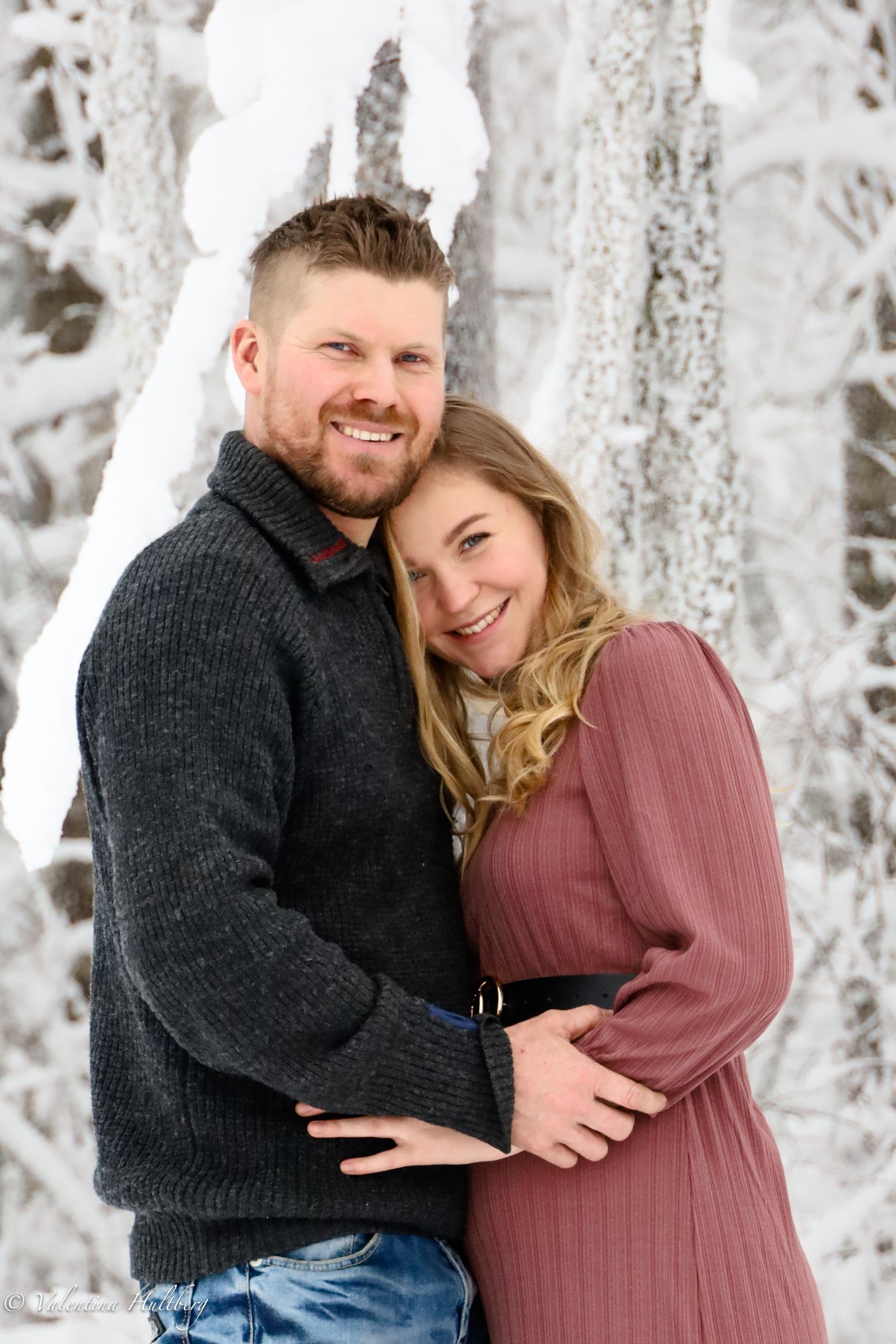 A couple embraces in a snowy winter forest setting wearing casual sweaters.