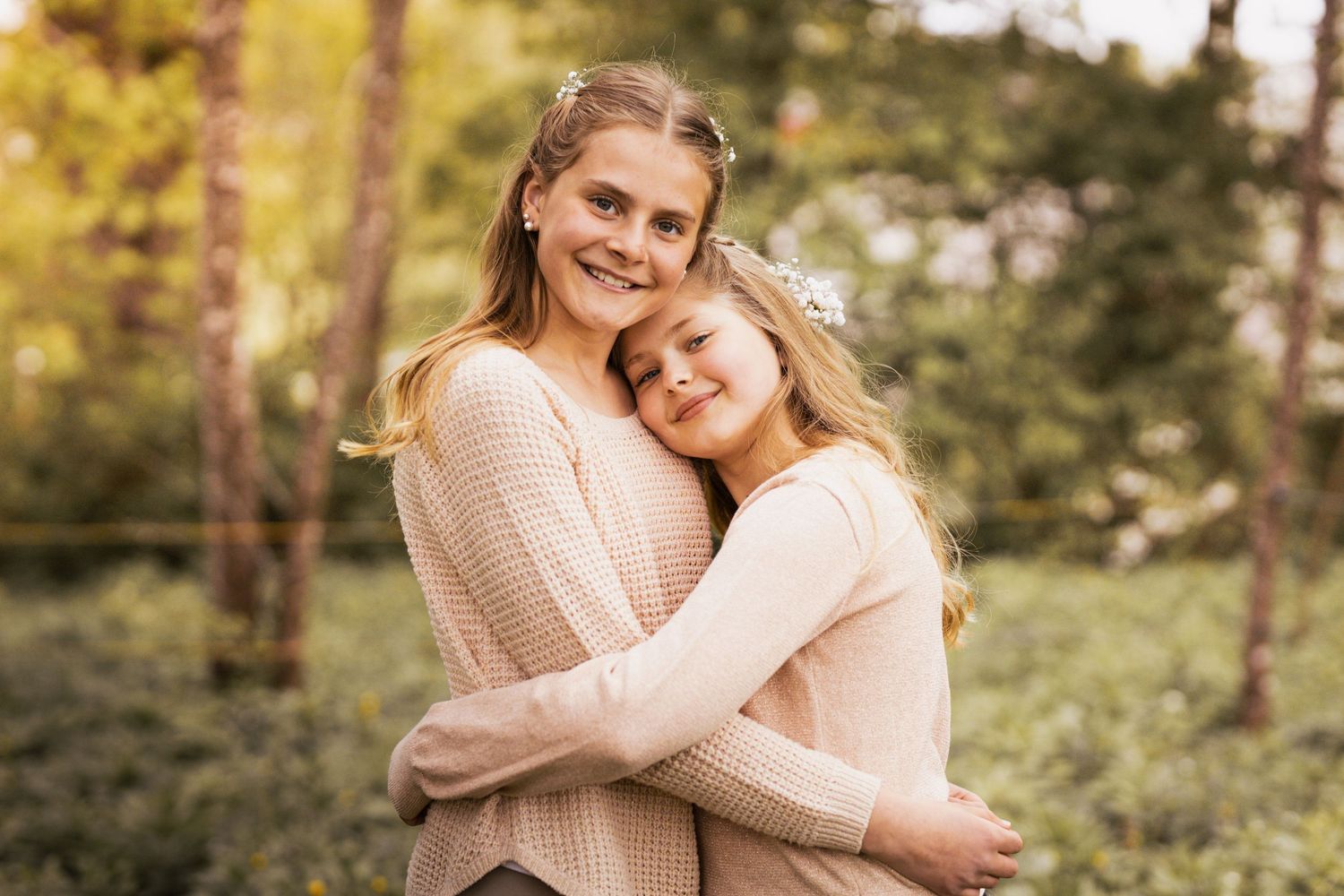 Two people in cream sweaters embrace while standing in a sunlit forest setting.