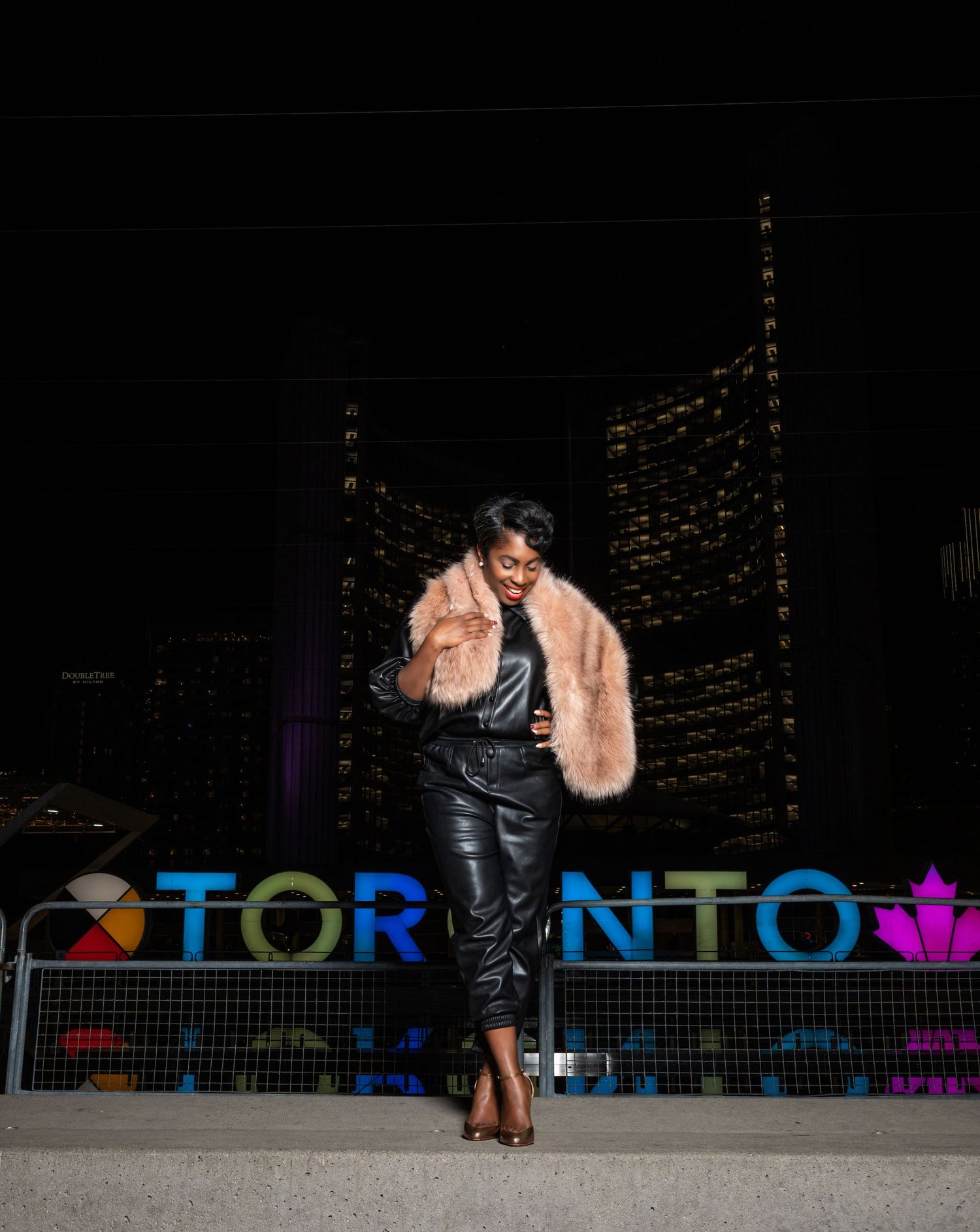 Woman in leather with a fur and heels at night in front of illuminated Toronto sign.