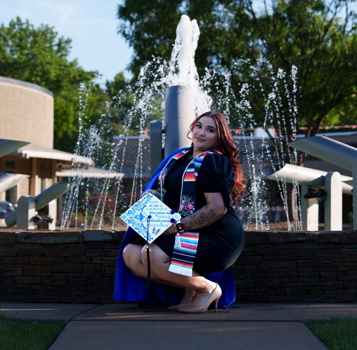A young woman in graduation attire poses by a fountain outdoors, holding a decorated cap and wearing a colorful stole.