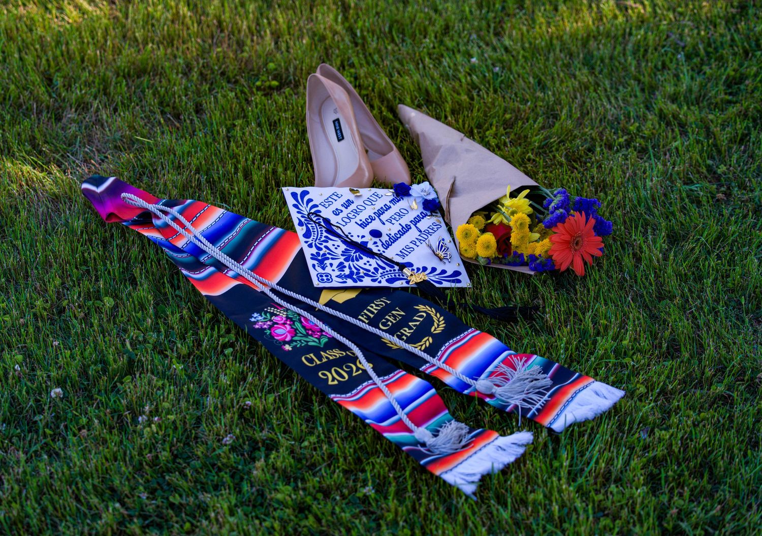 Graduation sash, pennant, diploma, flowers, and heels arranged on green grass for Class of 2023.