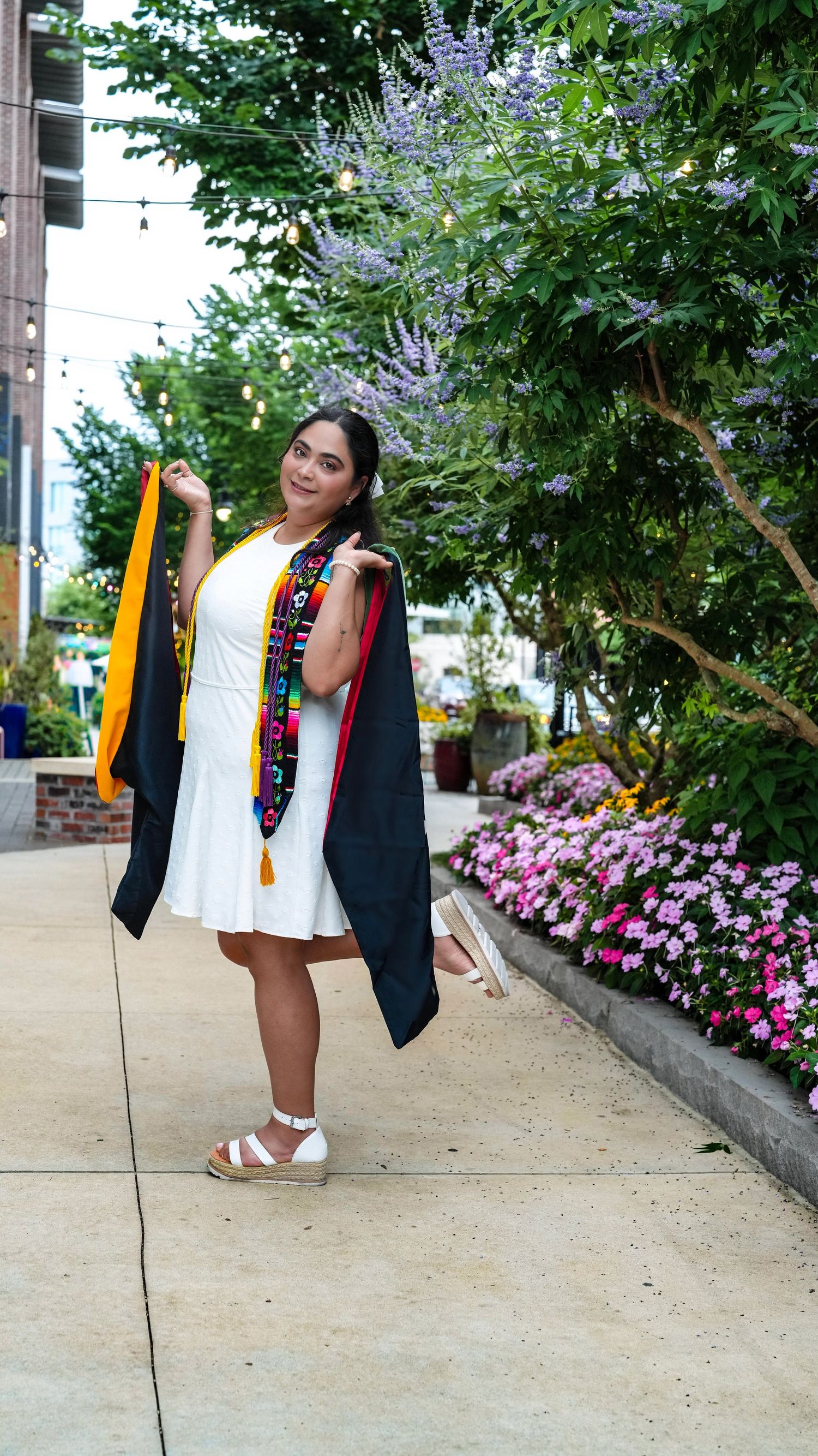 A smiling graduate poses in a black gown and colorful stole over a white dress, holding her cap outdoors near blooming flowers.