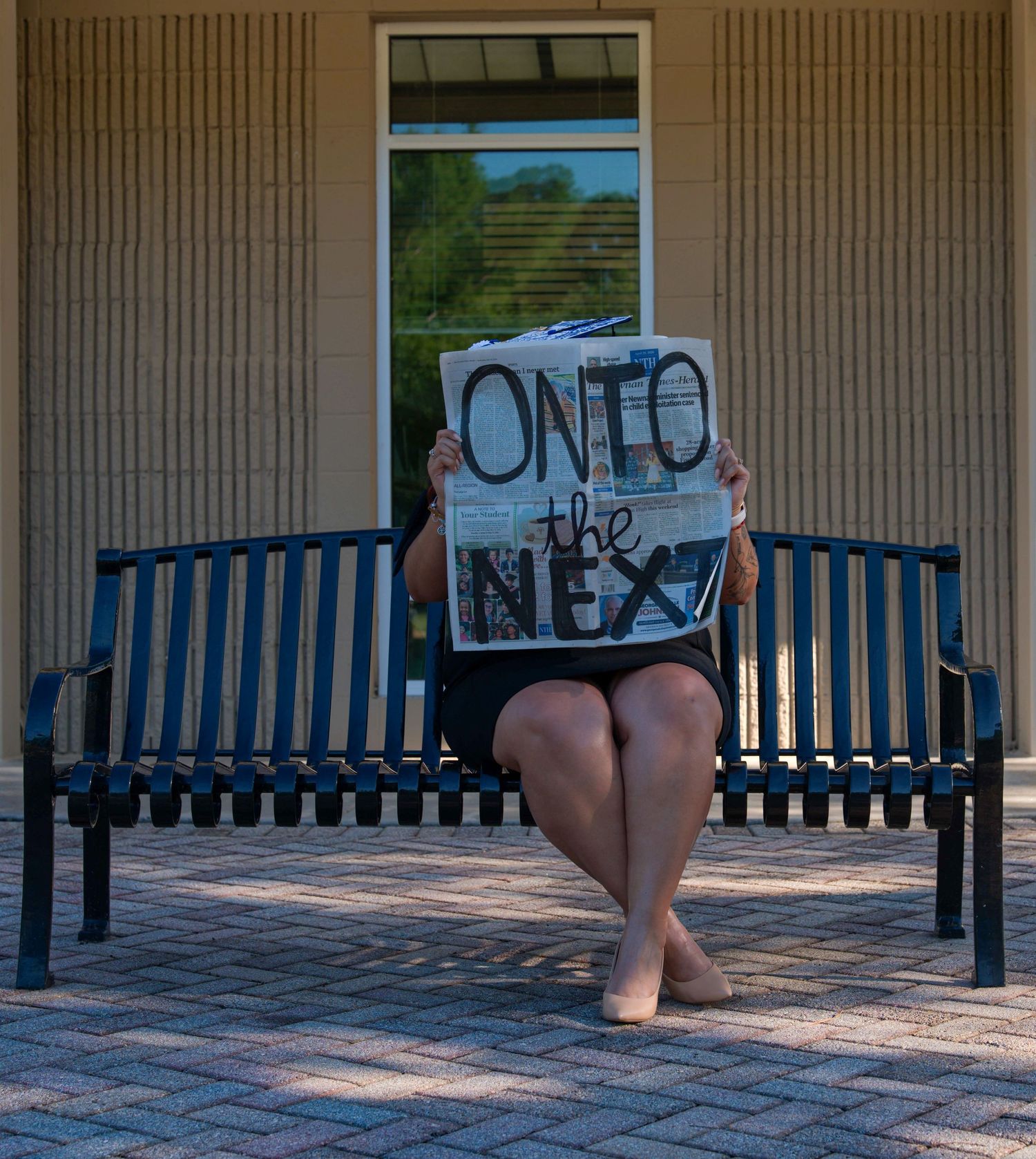 Person sitting on a bench holding a newspaper reading 'Onto the Next' outdoors near a brick building.