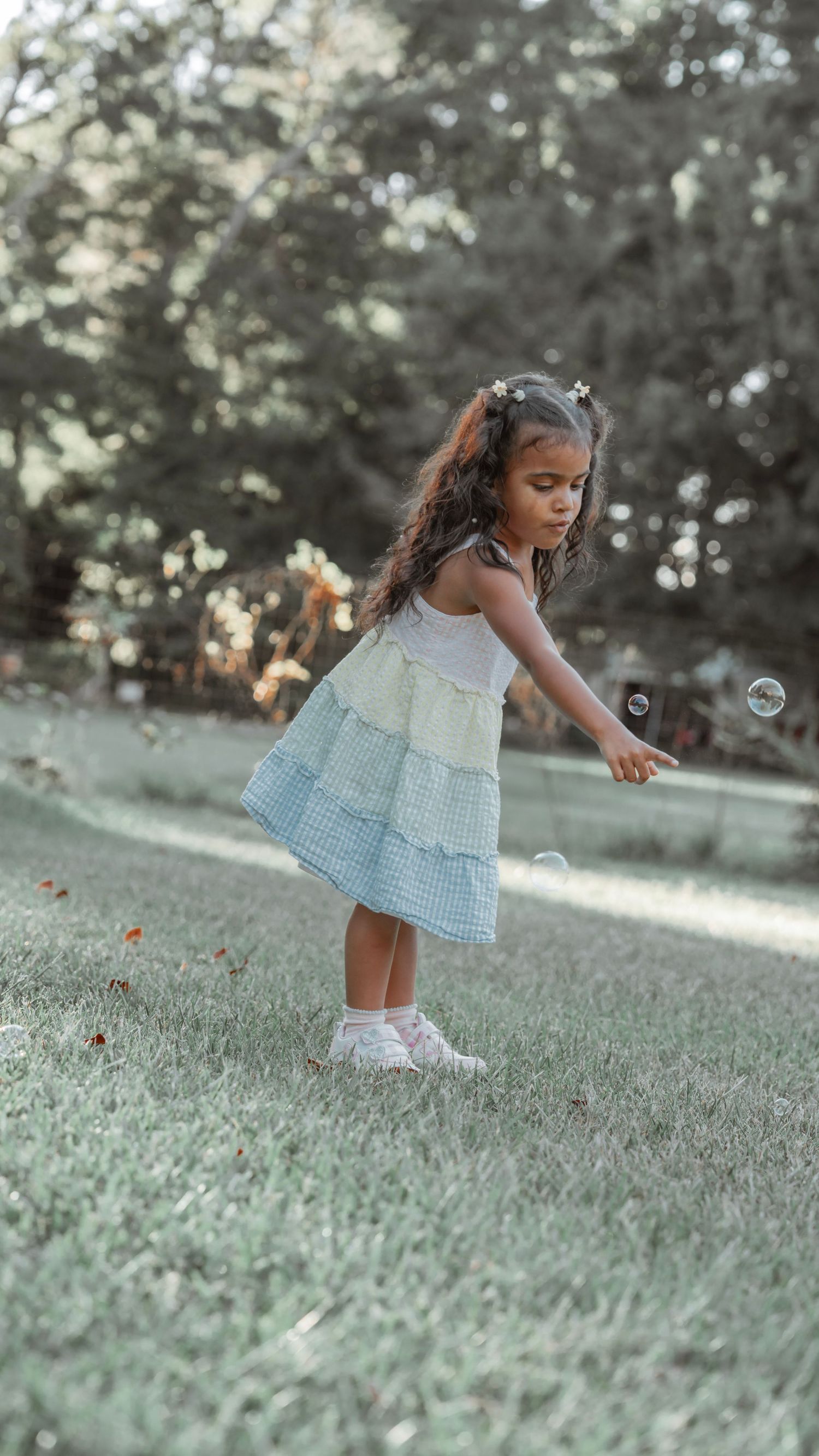 Young girl in tiered pastel dress reaching for bubbles in a sunlit green park.