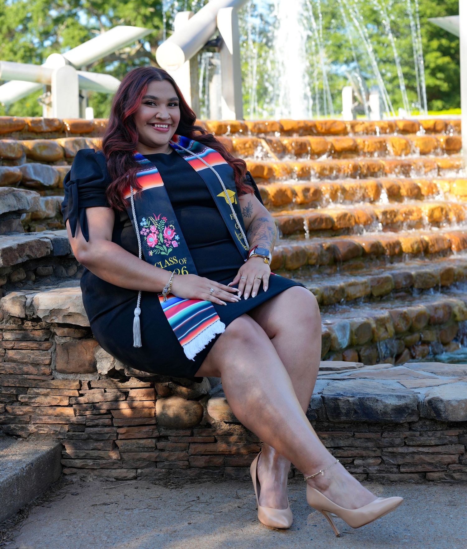 Woman in black dress with colorful sash sits smiling by a stone fountain outdoors.