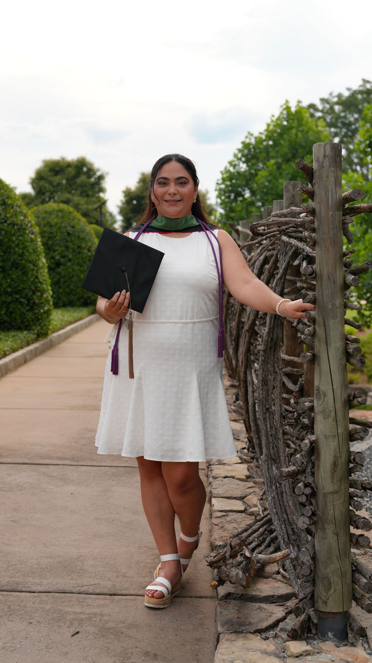 Young woman in white dress holding graduation cap and wearing academic regalia, posing outdoors on a garden path.