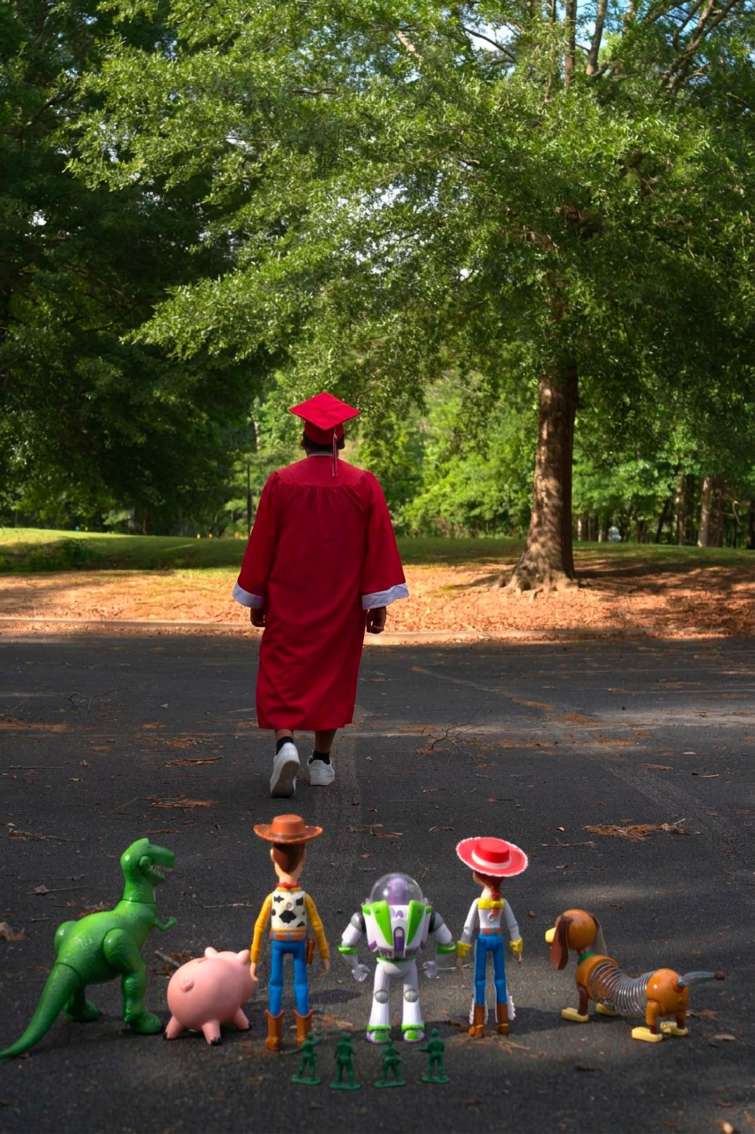 Graduate in red cap and gown walks away from Toy Story figurines including Woody, Buzz, and Rex on a park path.