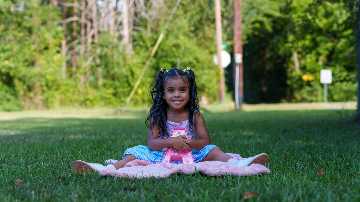 Young Black girl with braids sits cross-legged on a blanket in a lush green park, smiling at the camera.