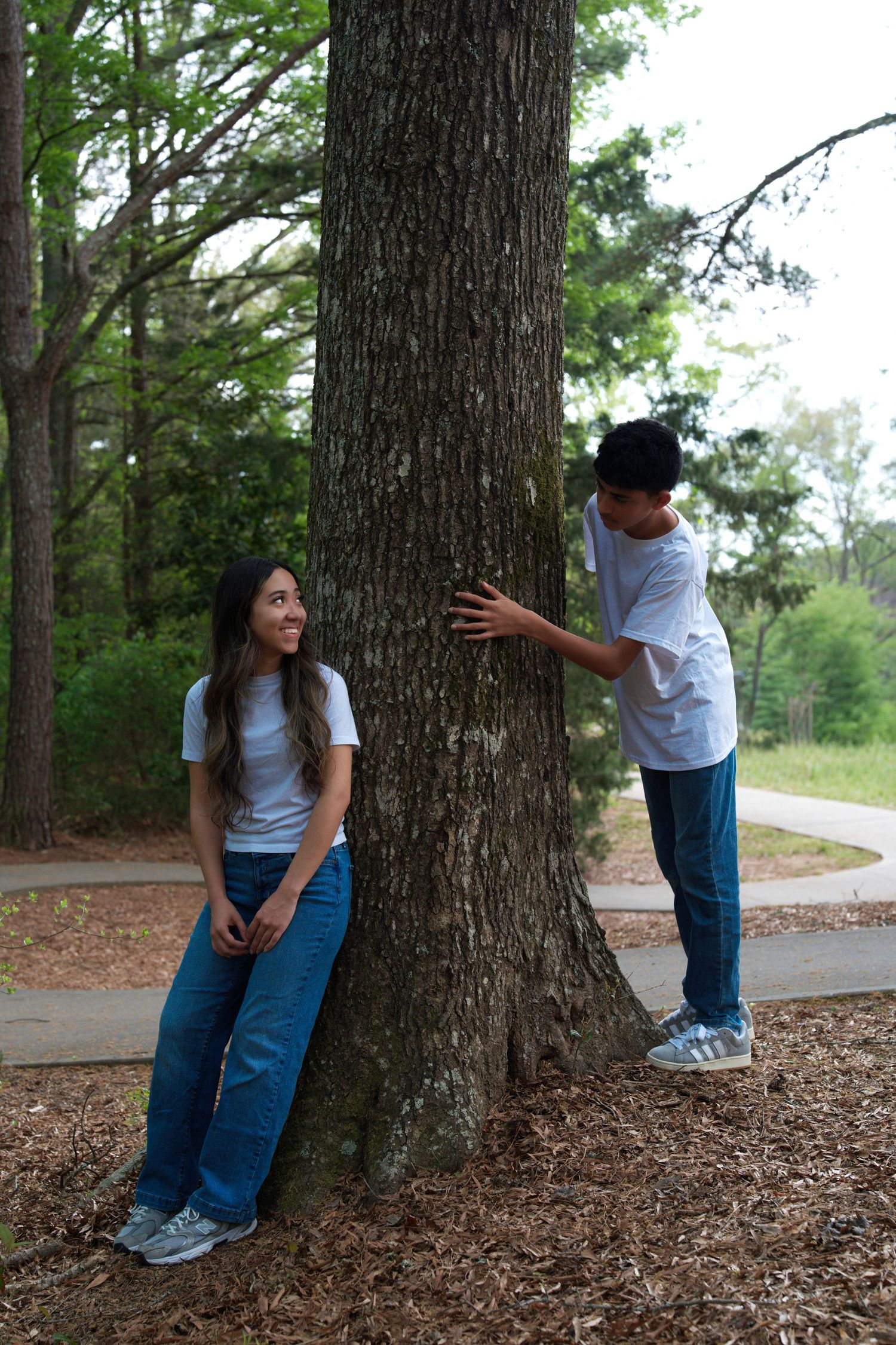 Two teens in white tees and jeans pose playfully by a large tree in a park setting.
