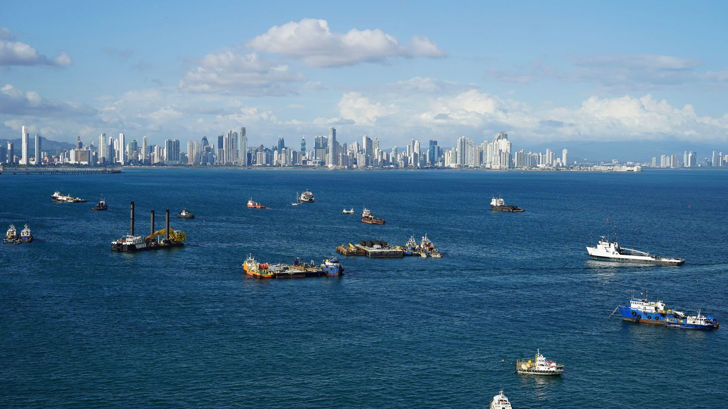 Panoramic view of Hong Kong's Victoria Harbor with modern skyscrapers and boats dotting the blue waters.