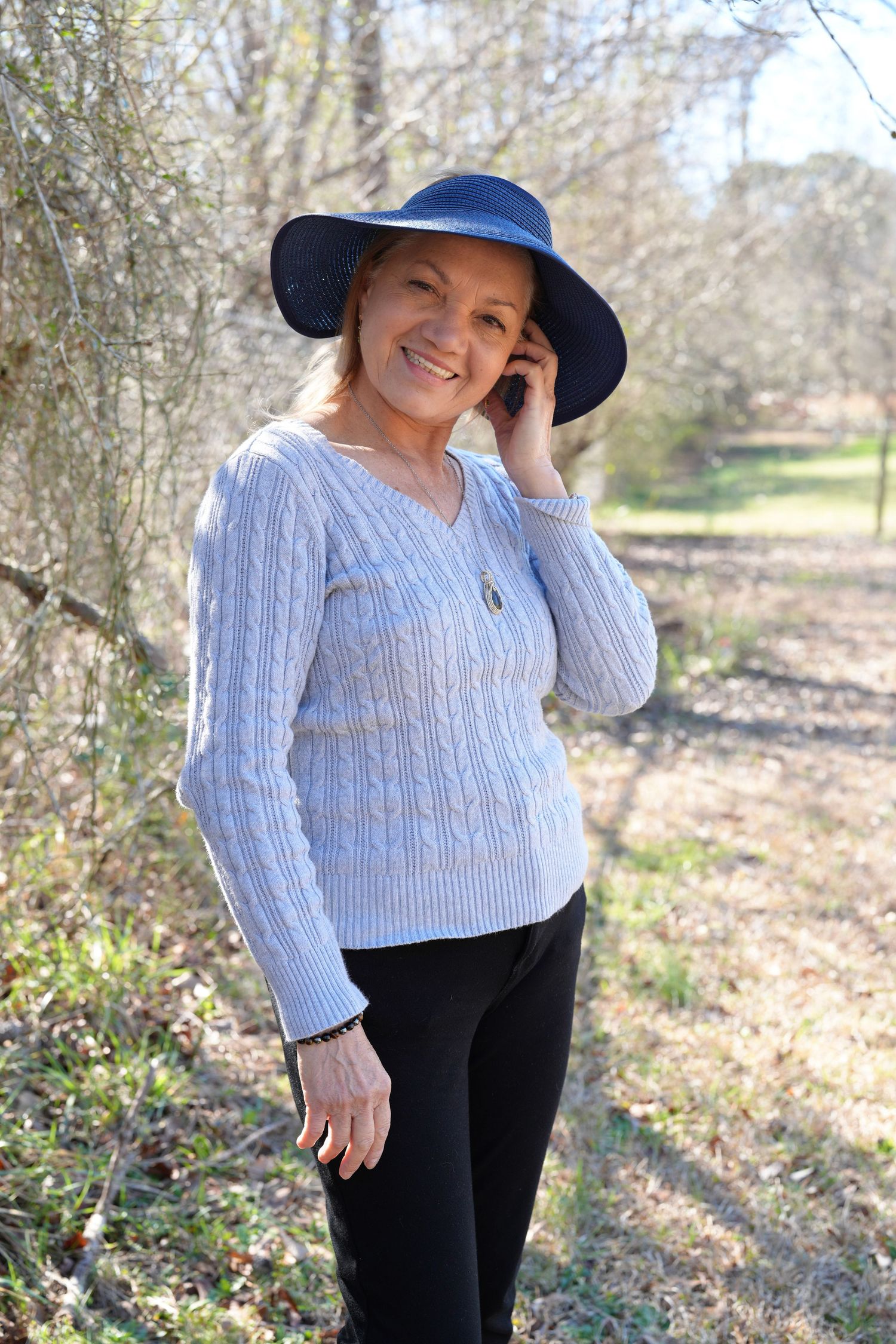 A person wearing a blue sunhat and striped sweater poses outdoors against a natural wooded background.