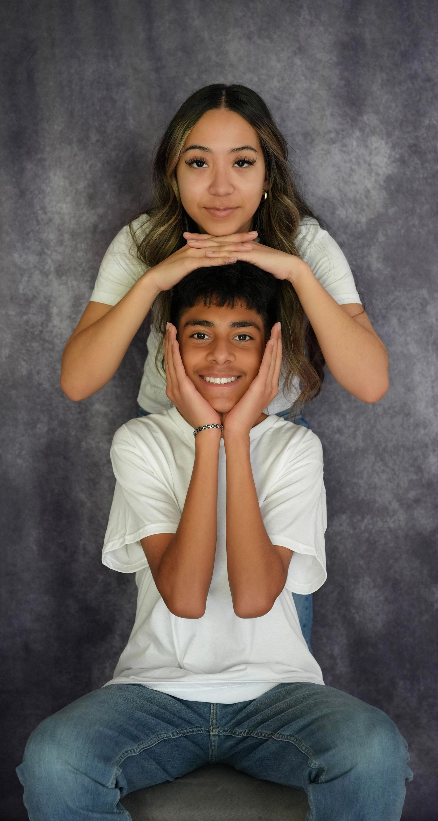Two smiling teens pose playfully in white shirts against a gray backdrop, the girl resting her chin on the boy's head.