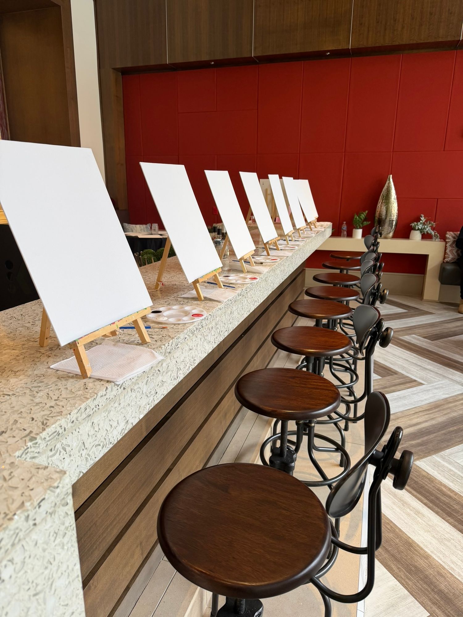 A row of blank canvases on easels along a granite counter with wooden bar stools, set up for a painting class.