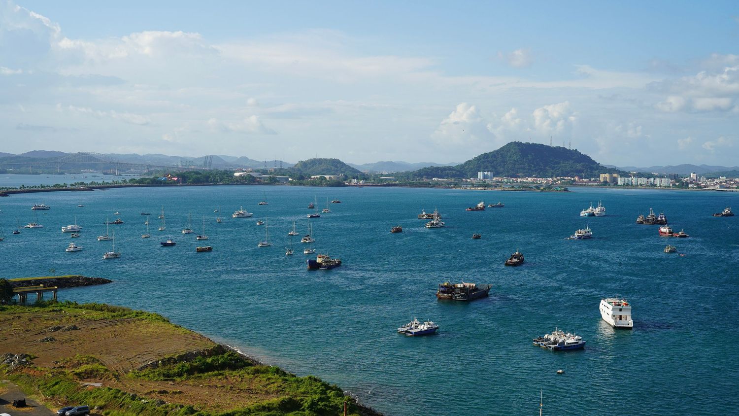 Panoramic view of a blue ocean bay with boats dotting the water and mountains in the distance.
