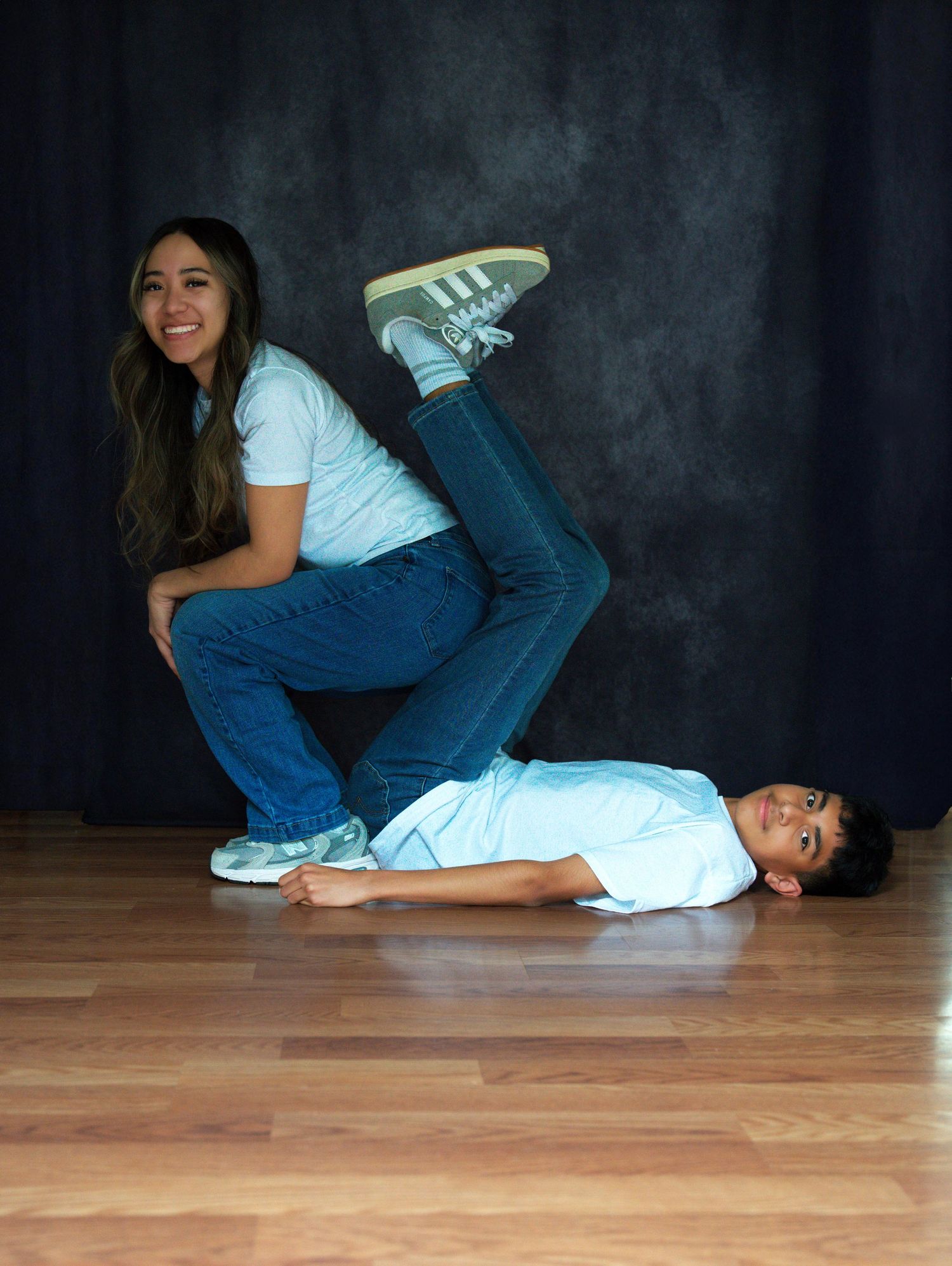 Two teens pose playfully, girl squatting while boy lies on floor with legs up in fun photo session.