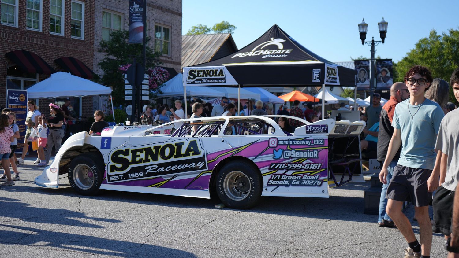Senola Raceway dirt track race car on display at an outdoor event with spectators and white tents in background.