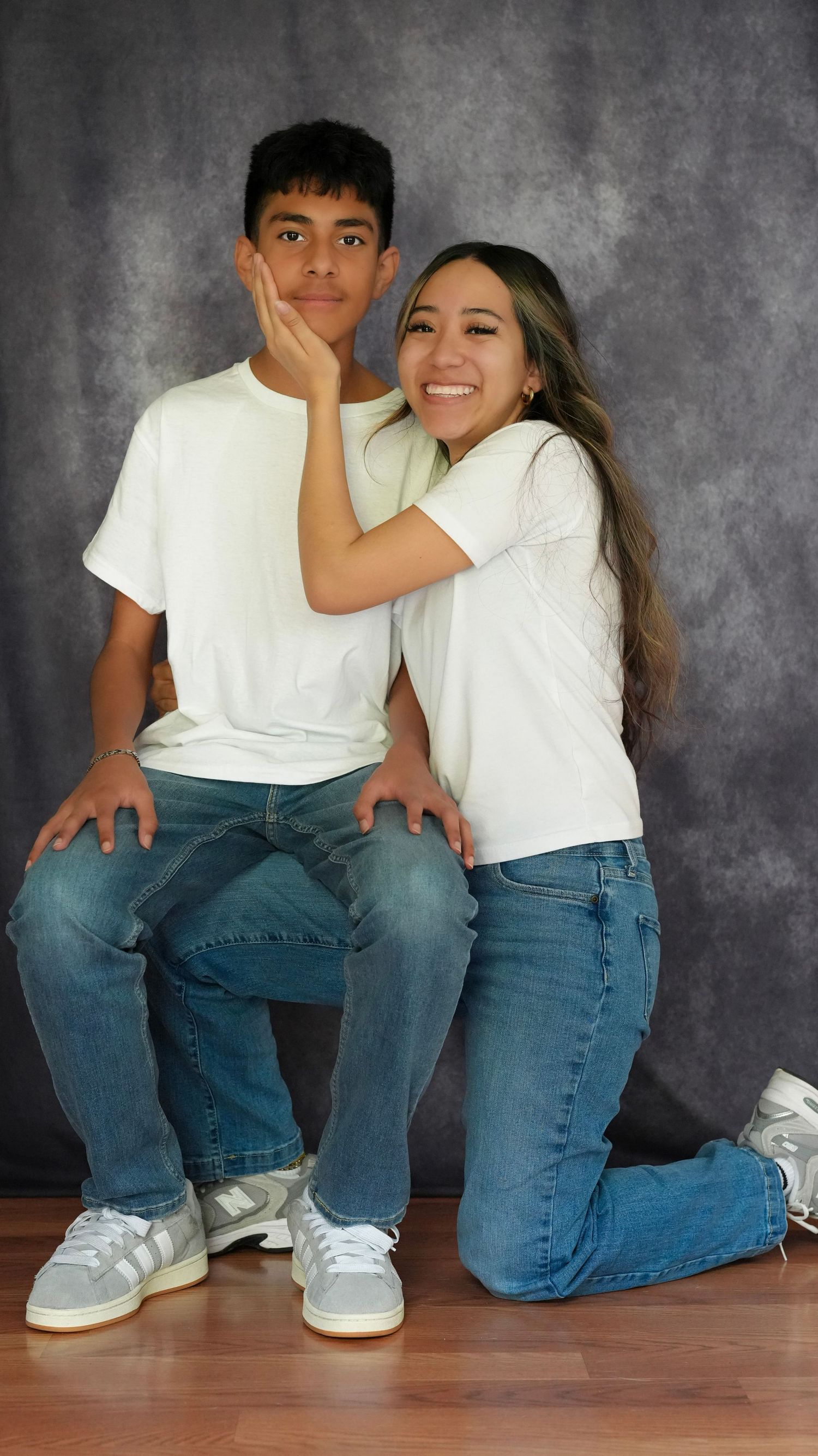 Two teens in matching white tees and jeans pose together smiling against a gray studio background.