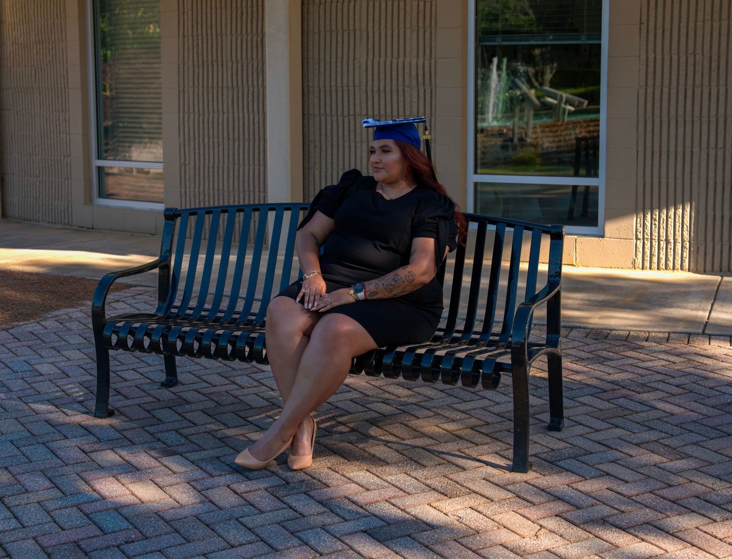 Graduate in cap and gown sitting on metal bench outdoors near brick building on sunny day.
