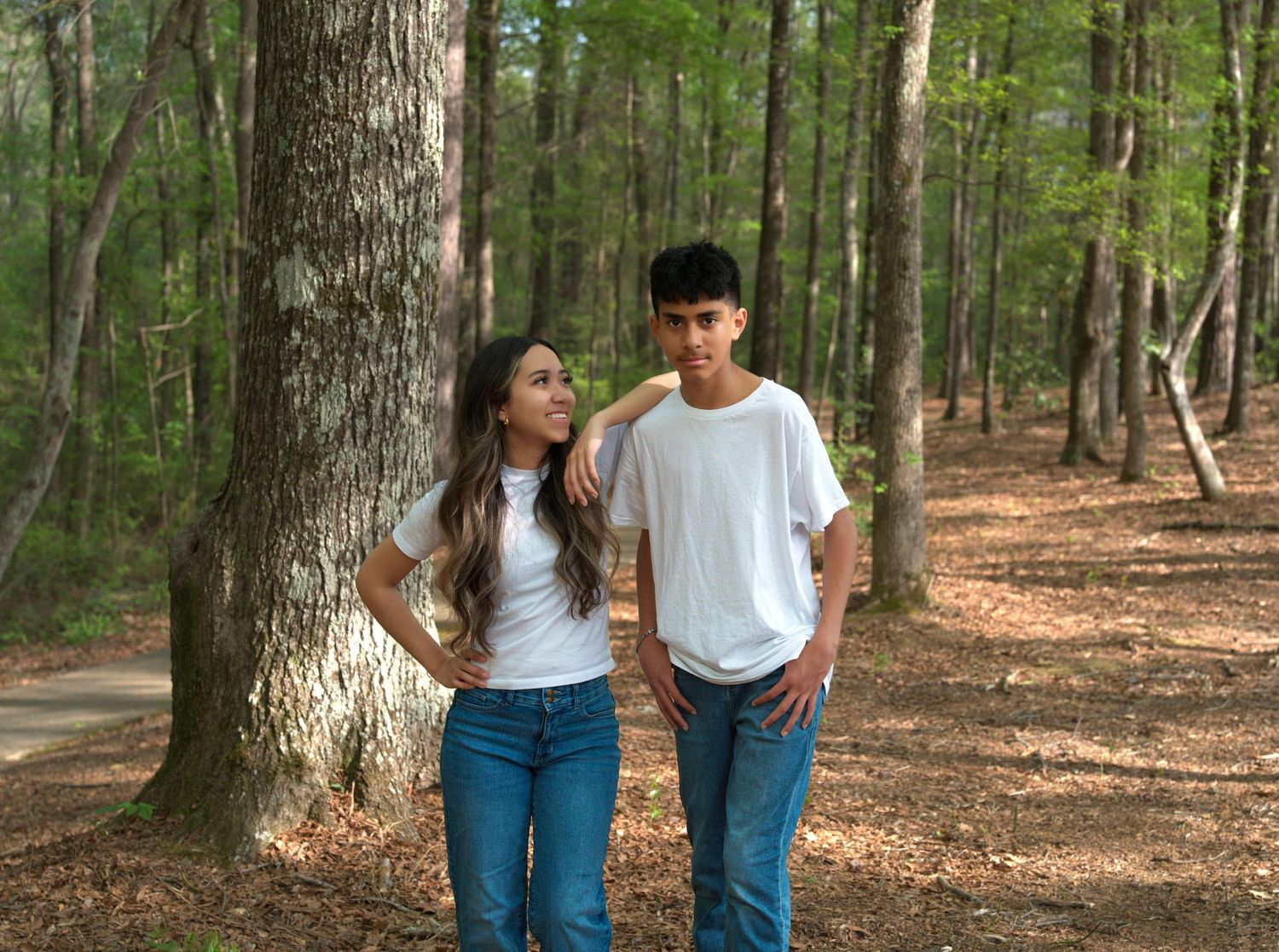 Two teens in white t-shirts and jeans walk together on a forest path surrounded by tall trees.