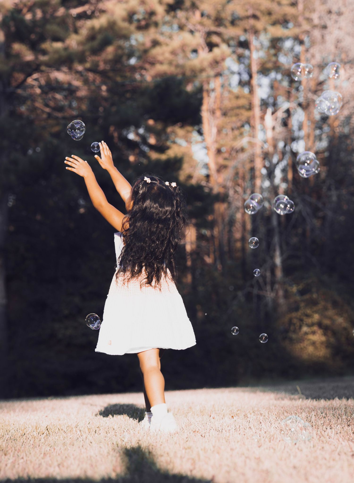 A young girl in a white dress reaches for floating soap bubbles in a sunlit forest clearing.