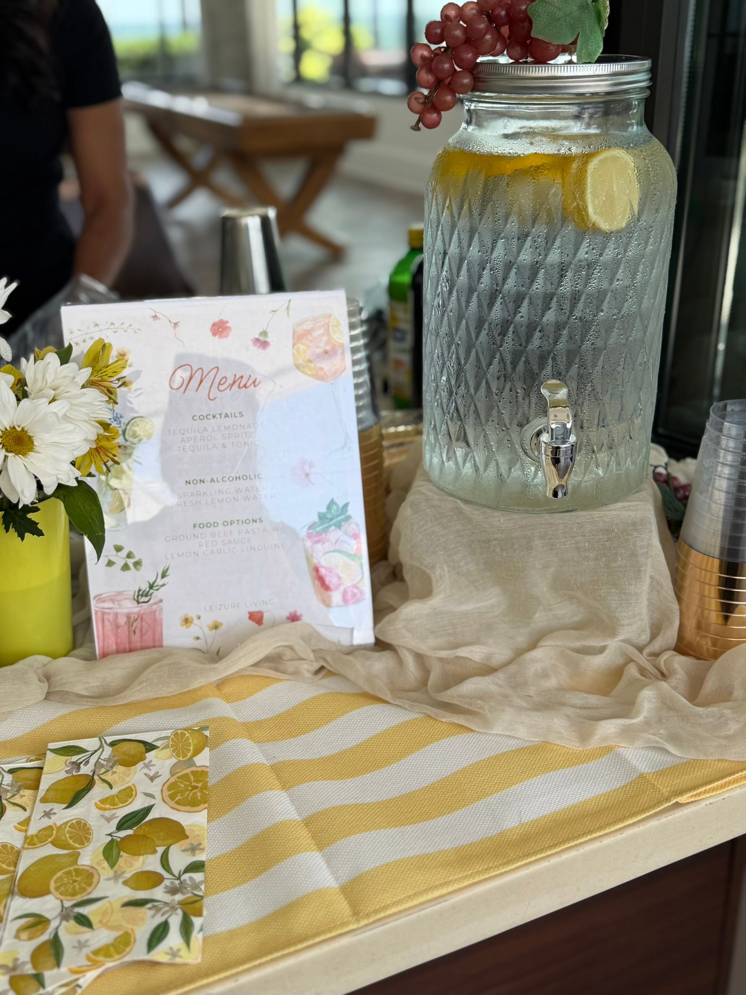 Elegant beverage dispenser with lemon water, floral menu card, white daisies, and lemon-print napkins on a decorated party table.