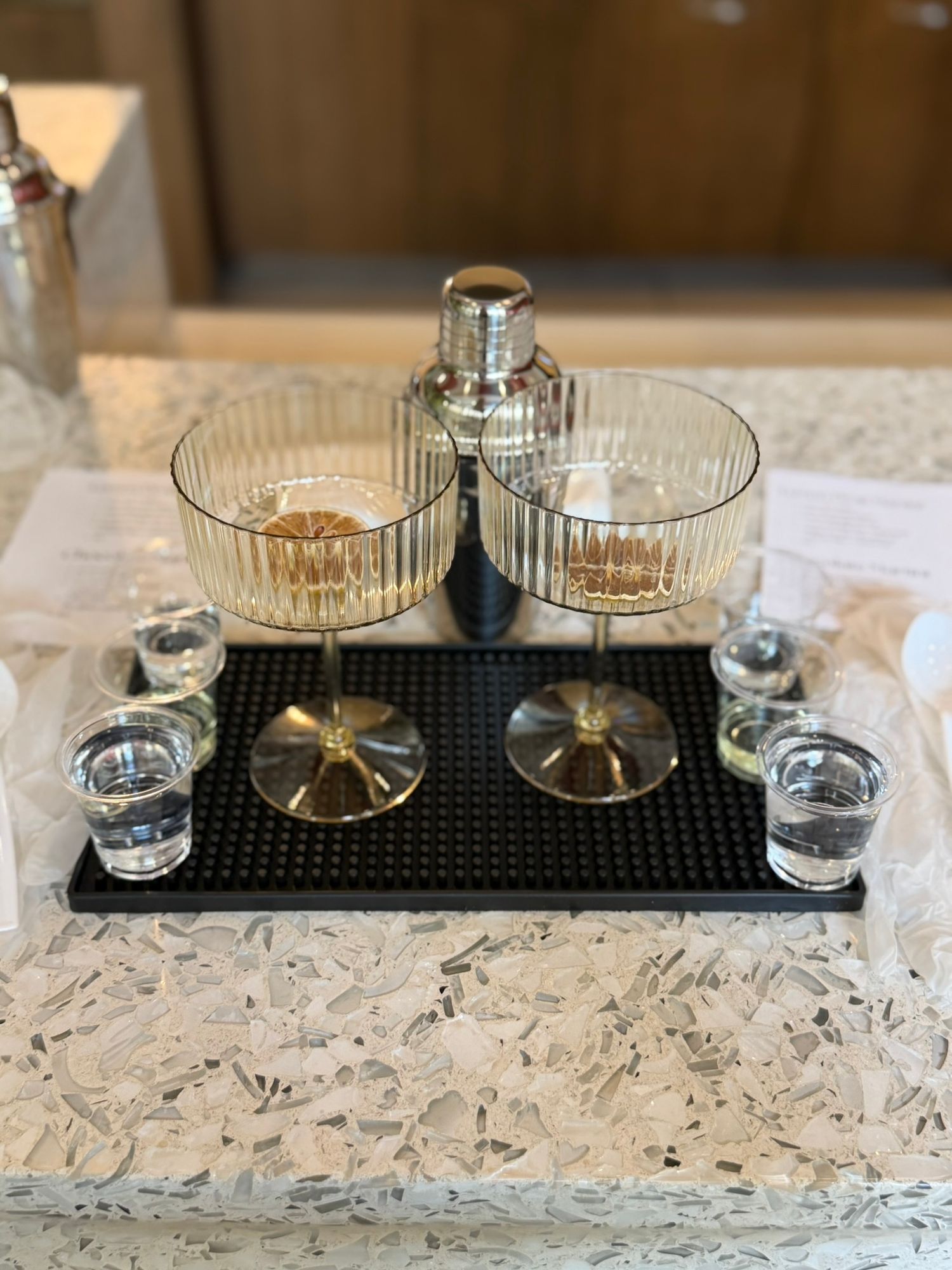 Two elegant ribbed coupe cocktail glasses with gold bases, shaker, and shot glasses on a black bar mat.